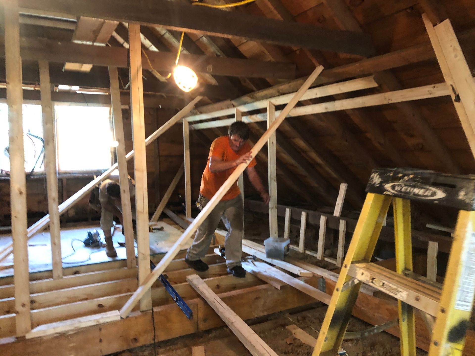 Workers framing an attic, with exposed beams, ladders, and tools in a dim unfinished room.