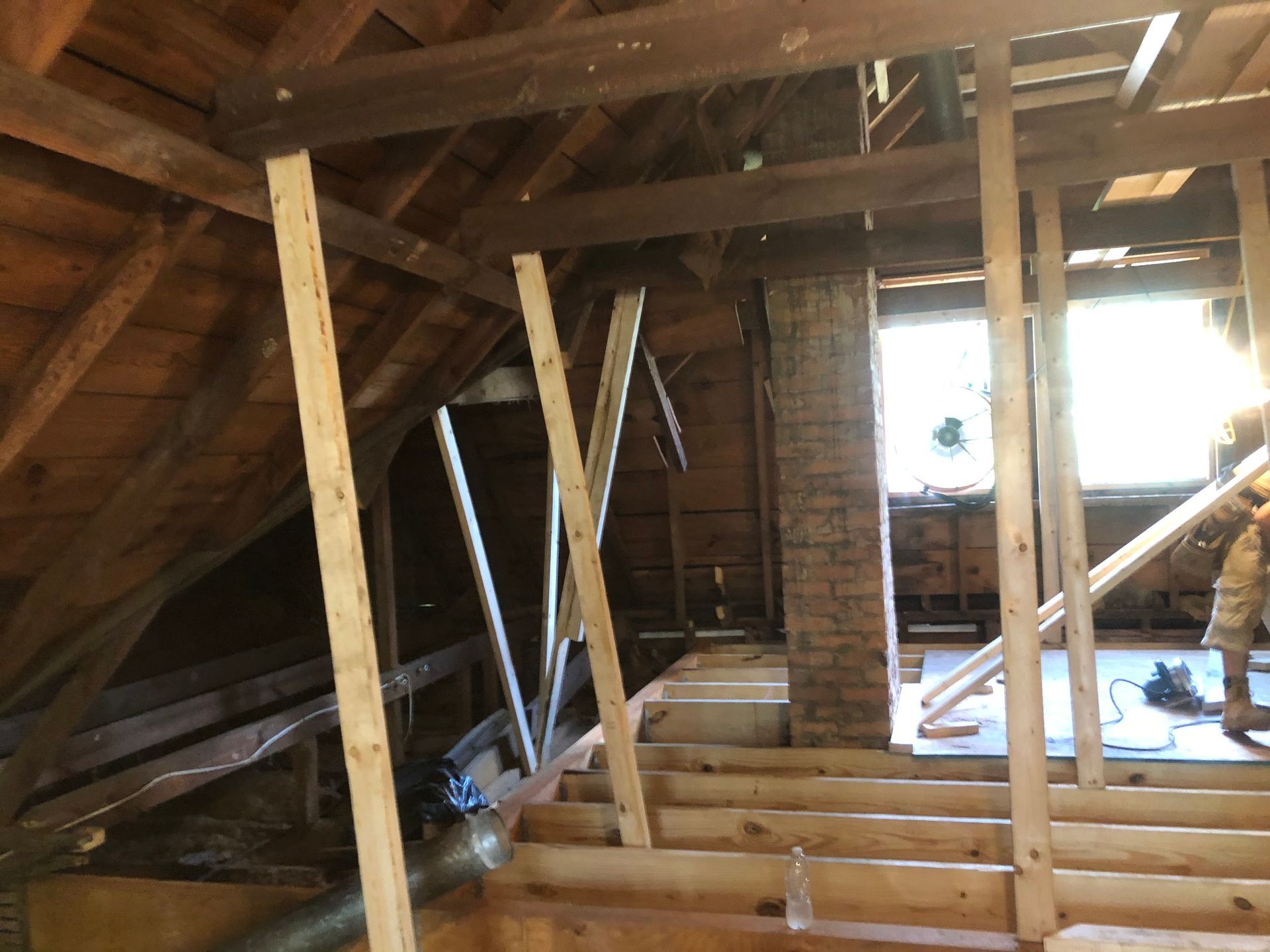 Interior of an unfinished attic with exposed wooden beams, framing, and construction materials near a bright window.