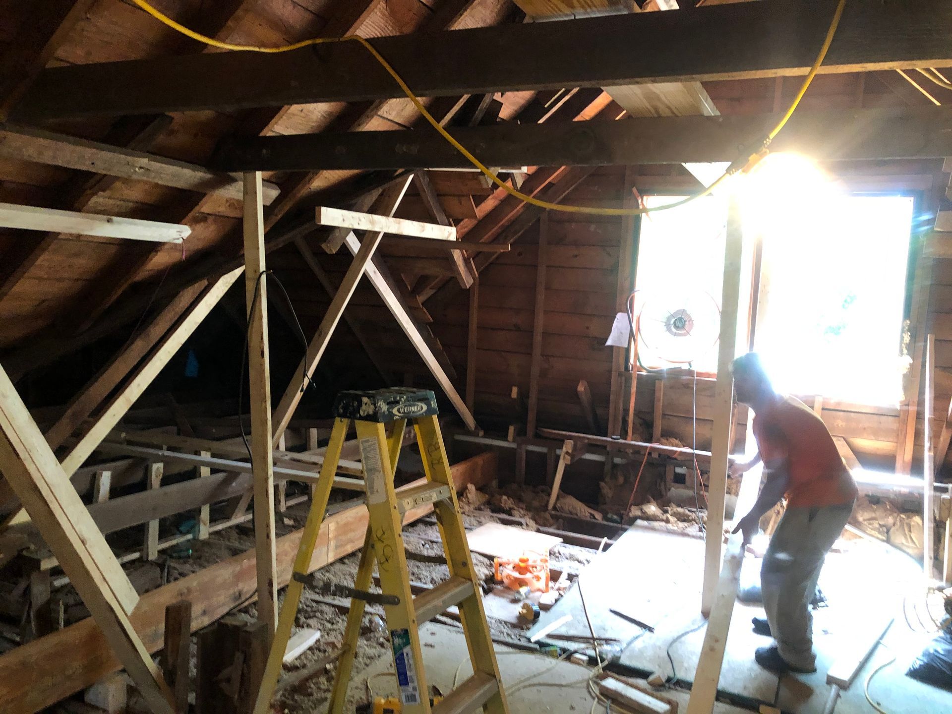 Worker in a dusty attic beside a ladder and bright window, cleaning debris from the floor.