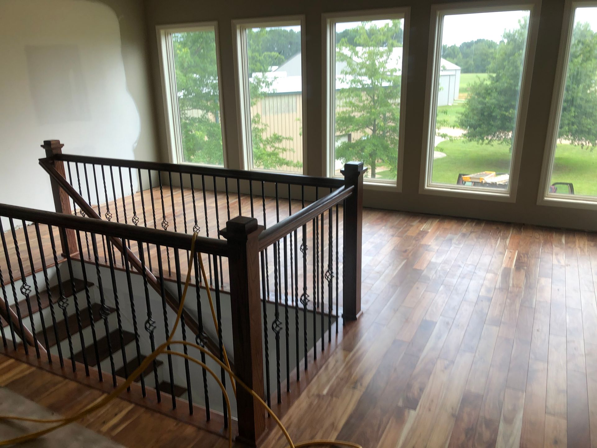 Bright empty landing with stairs, black railing, hardwood floors, and large windows overlooking trees.