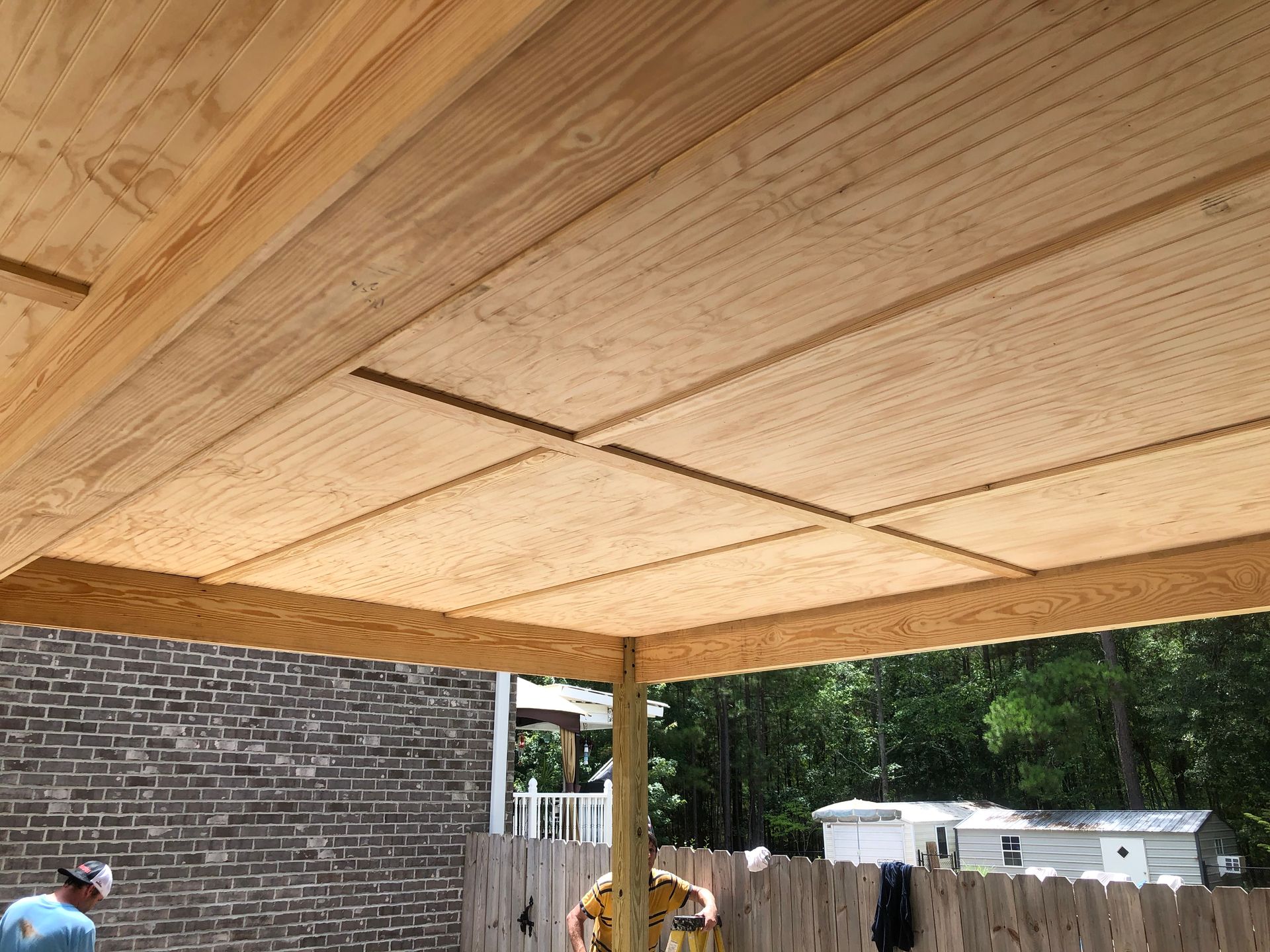 Wooden patio roof under construction, with exposed beams and a support post beside a brick house.