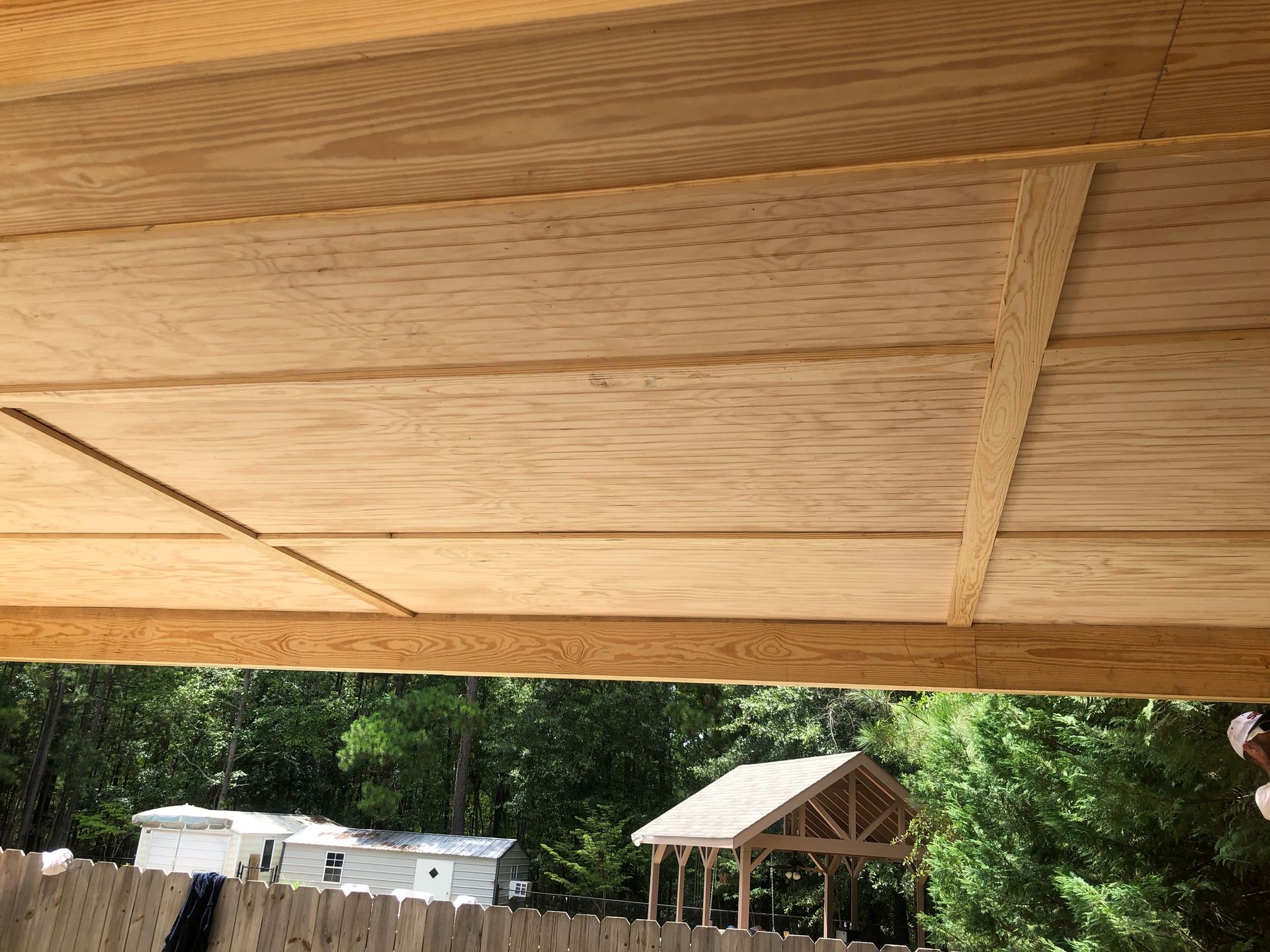 Wooden pavilion roof over a wooded outdoor area with a small structure in the background