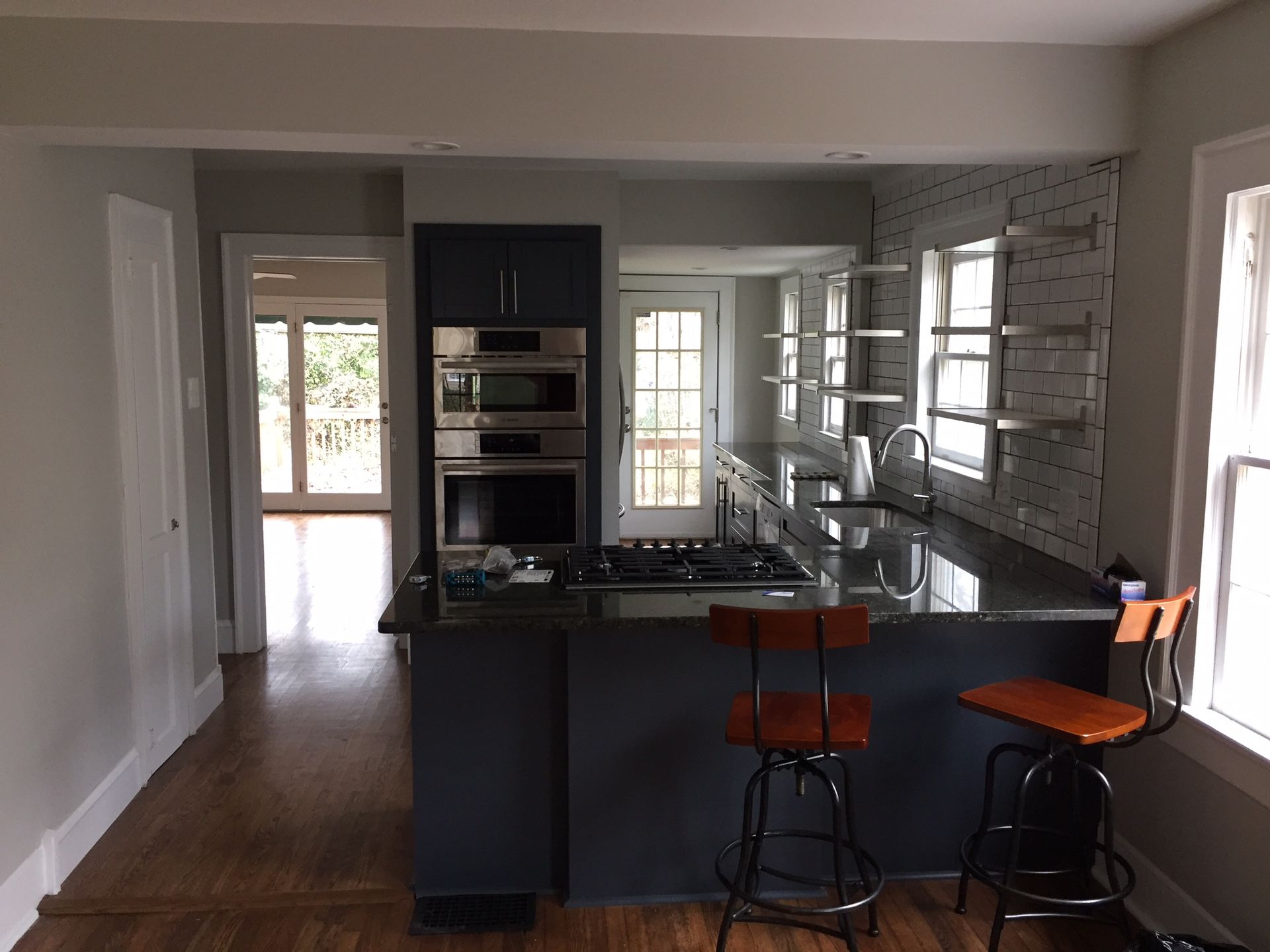 Modern kitchen with dark island, orange stools, and glass-front cabinets; doorway to a bright room.