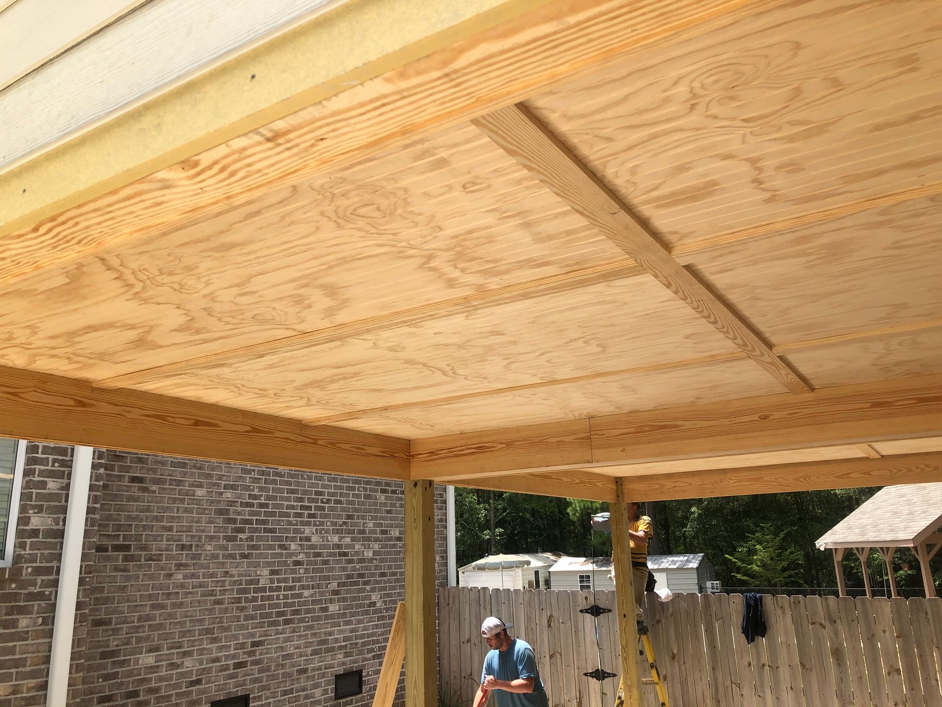 Wooden patio roof under construction beside a brick house, with a worker visible below.