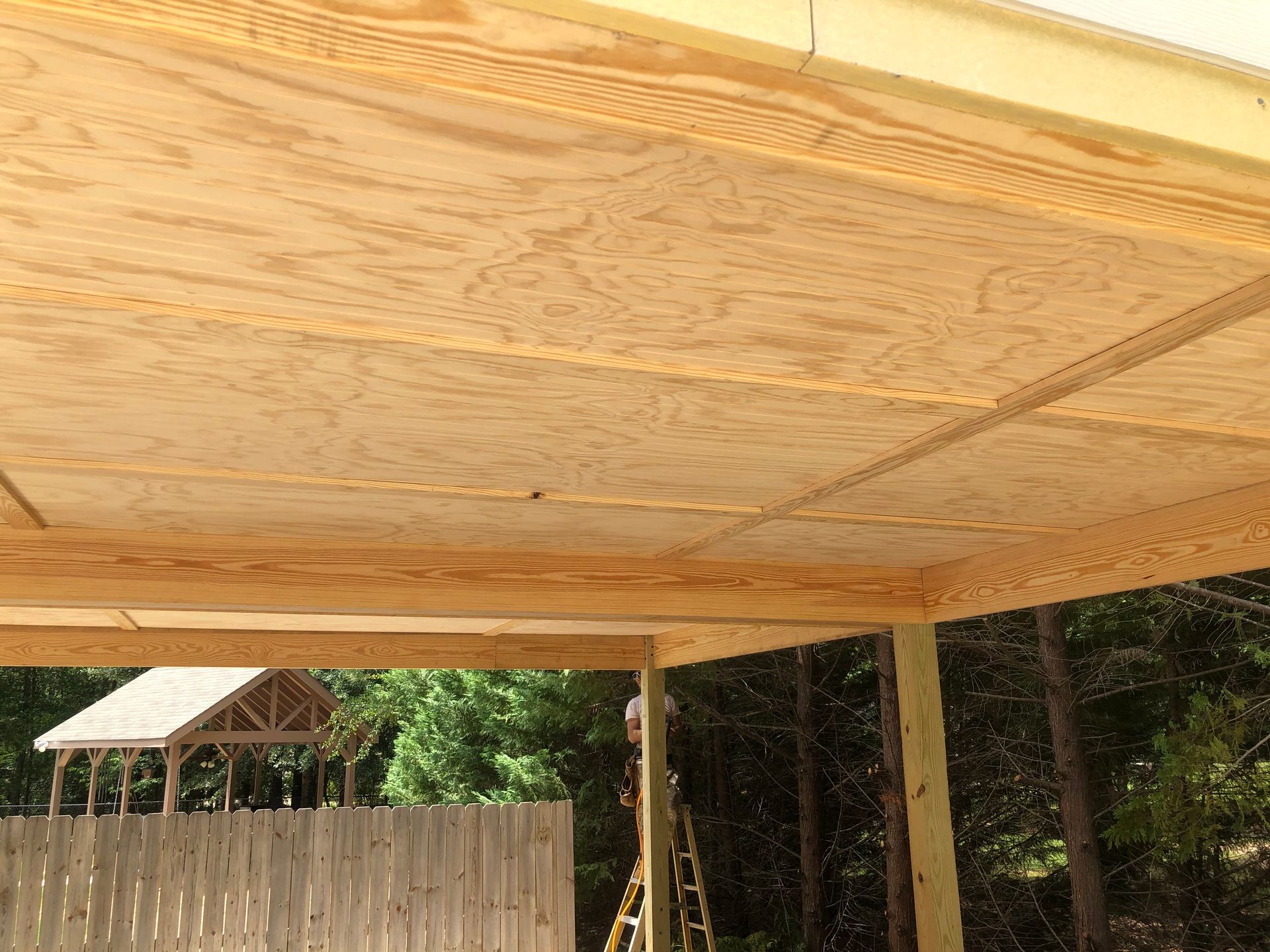 Wooden canopy ceiling over a fenced backyard with trees in the background.