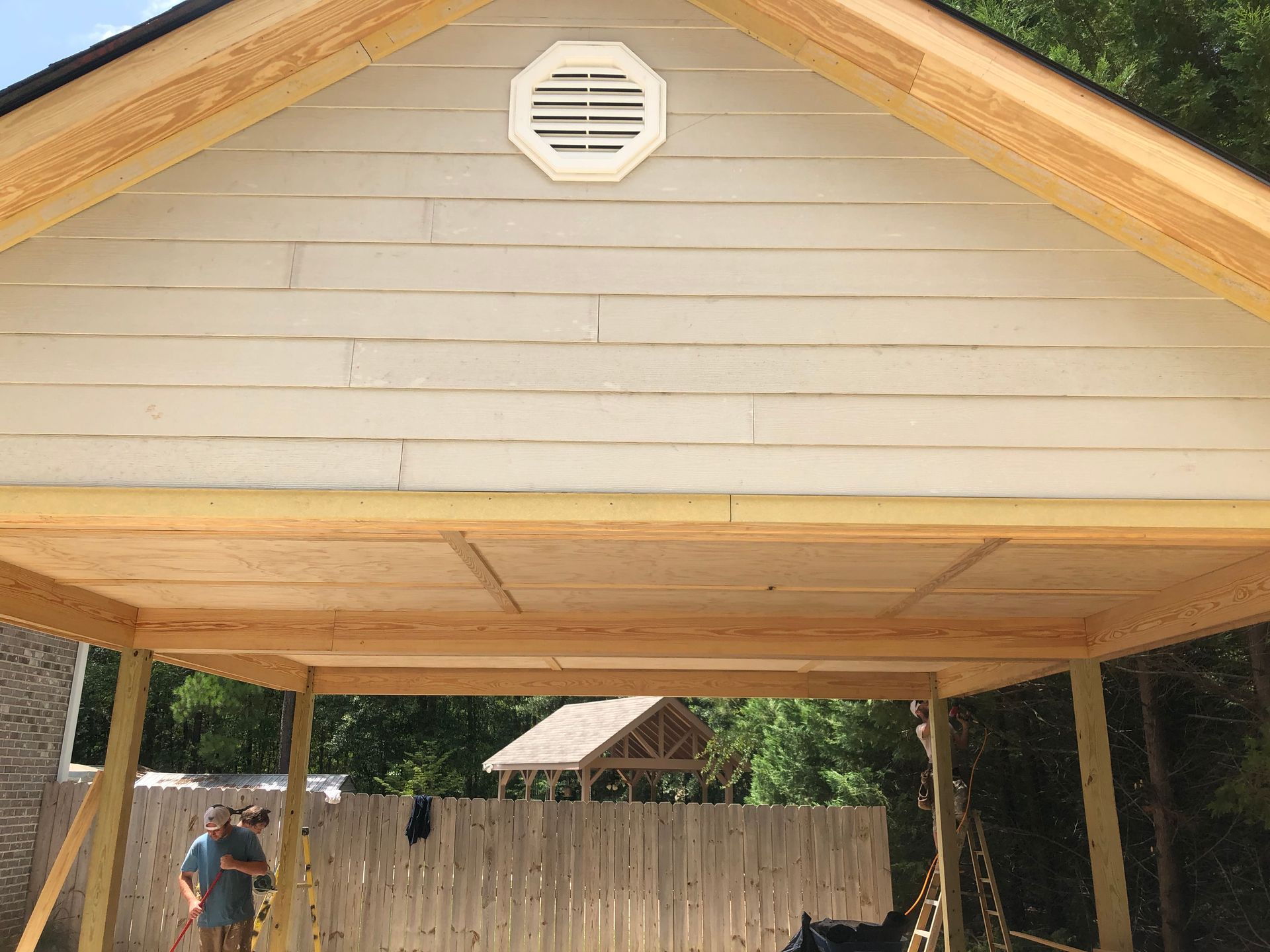 Covered wooden carport with a peaked roof and one person standing underneath near the left side.