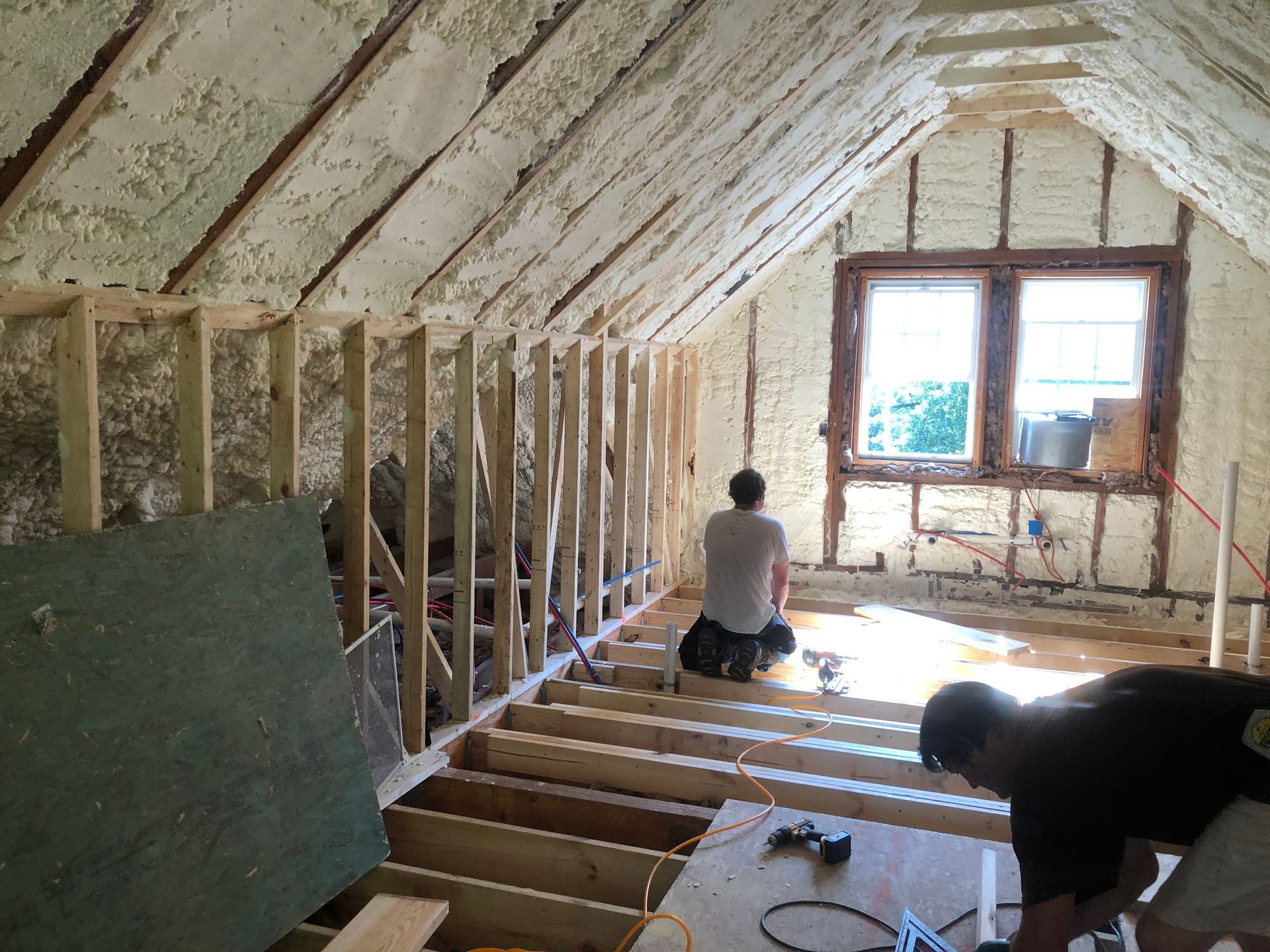 Workers installing insulation in an unfinished attic near a window.