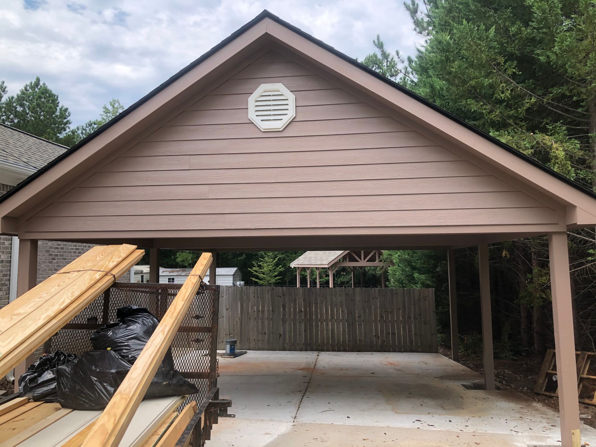 Brown carport with gabled roof and wooden ramps, concrete floor, and a fenced backyard behind it