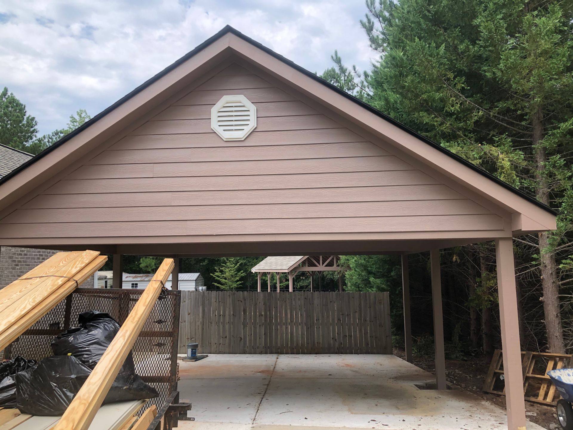 Carport with a tan gable roof and wooden posts, viewed from the driveway, with trees in the background