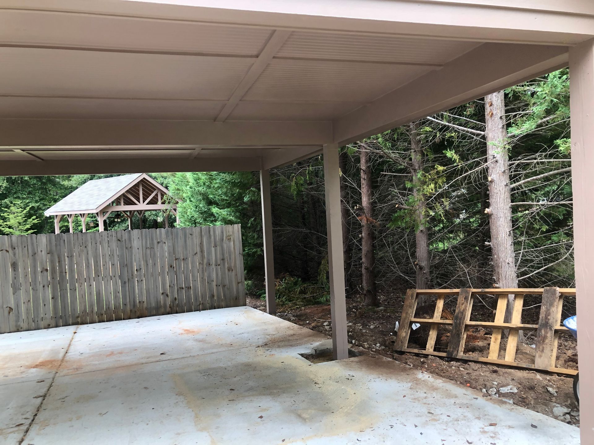 Covered carport with concrete floor, wooden fence, and stacked pallets along the right side