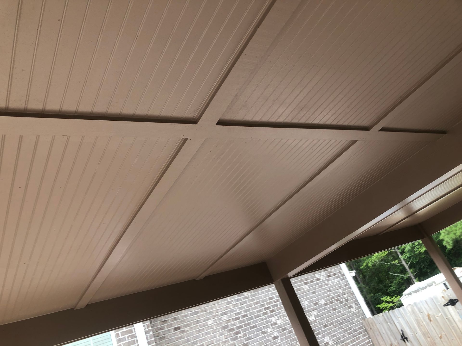 Beige patio ceiling panels above a shaded outdoor area with a sliver of sky and trees at the edge