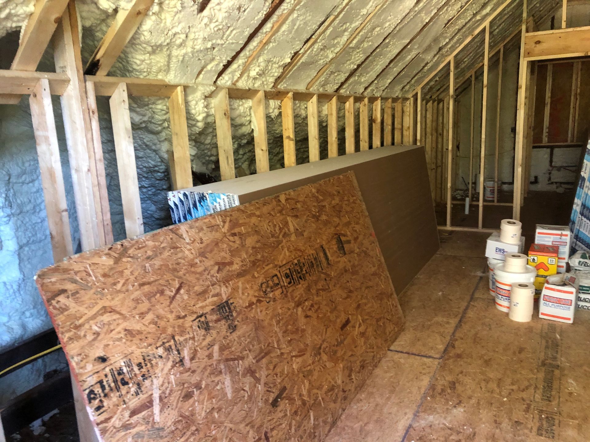 Unfinished attic with exposed insulation, plywood sheets, and paint cans on the floor.