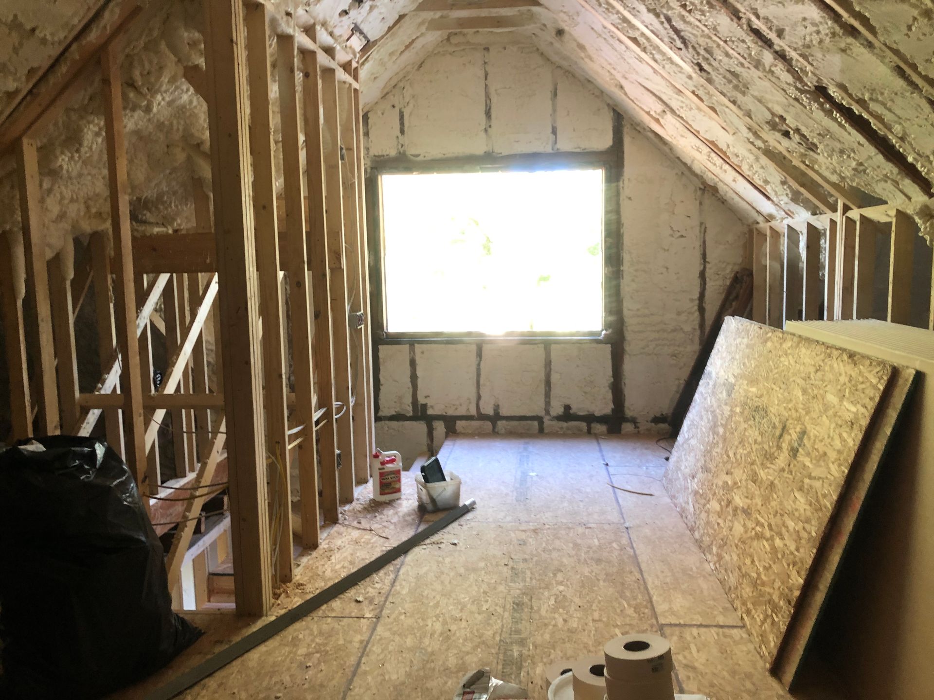 Unfinished attic room with exposed wall studs, plywood floor, insulation boards, and a bright window at the end.