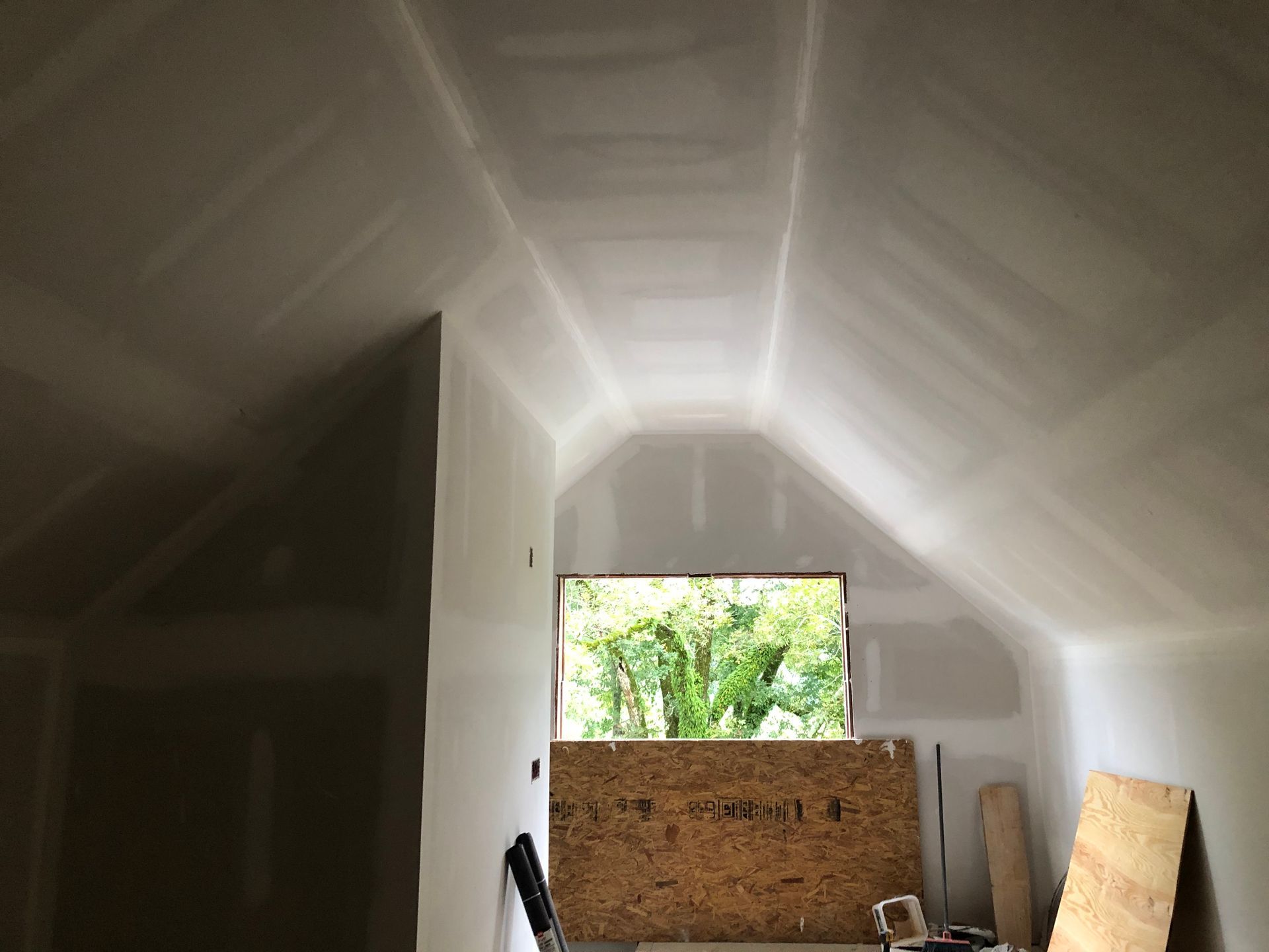 Interior of an unfinished attic with white insulation, exposed wood framing, and a window opening to greenery.