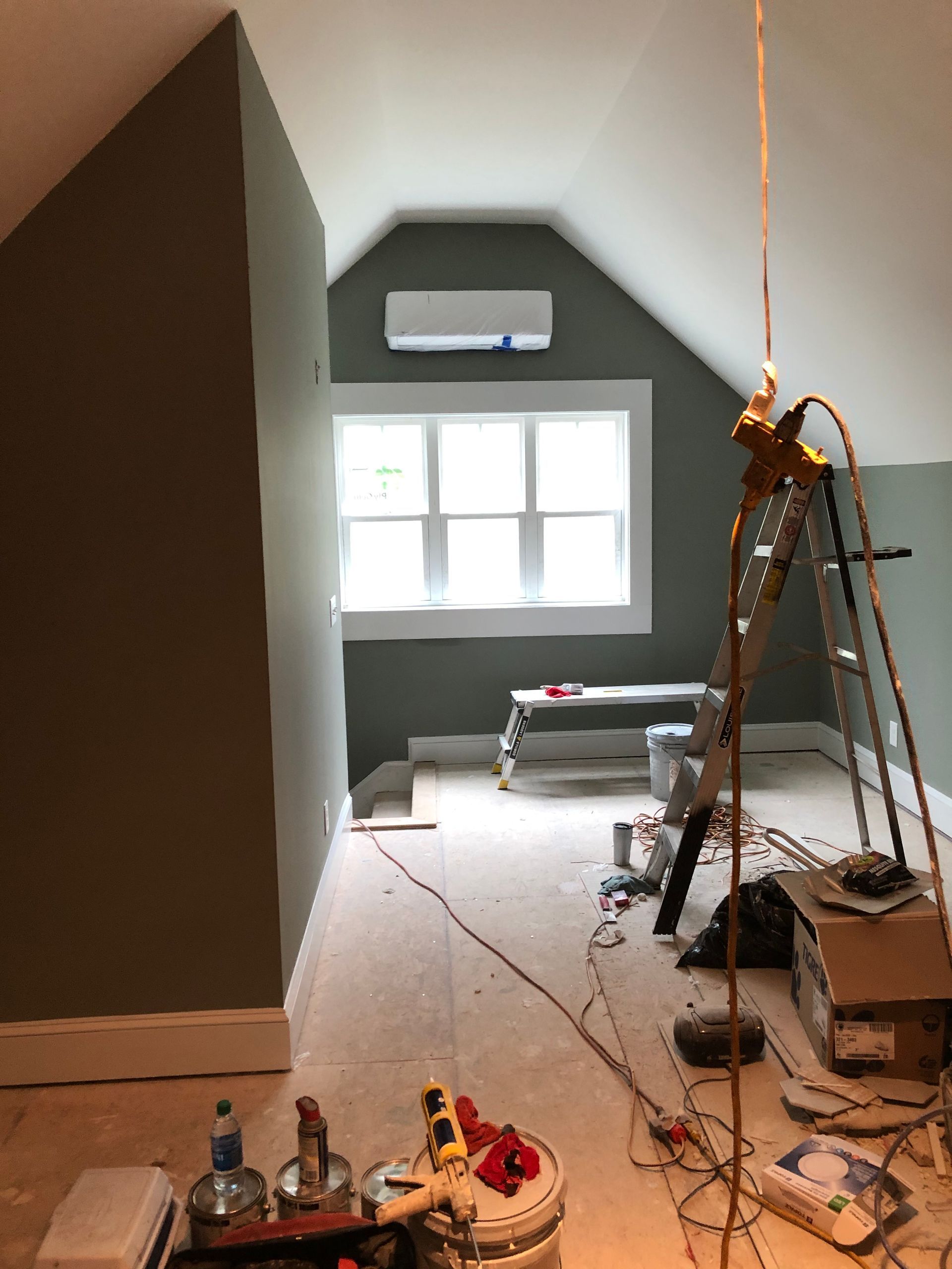 Narrow attic room under renovation with window, wall-mounted AC, ladder, tools, and debris on the floor