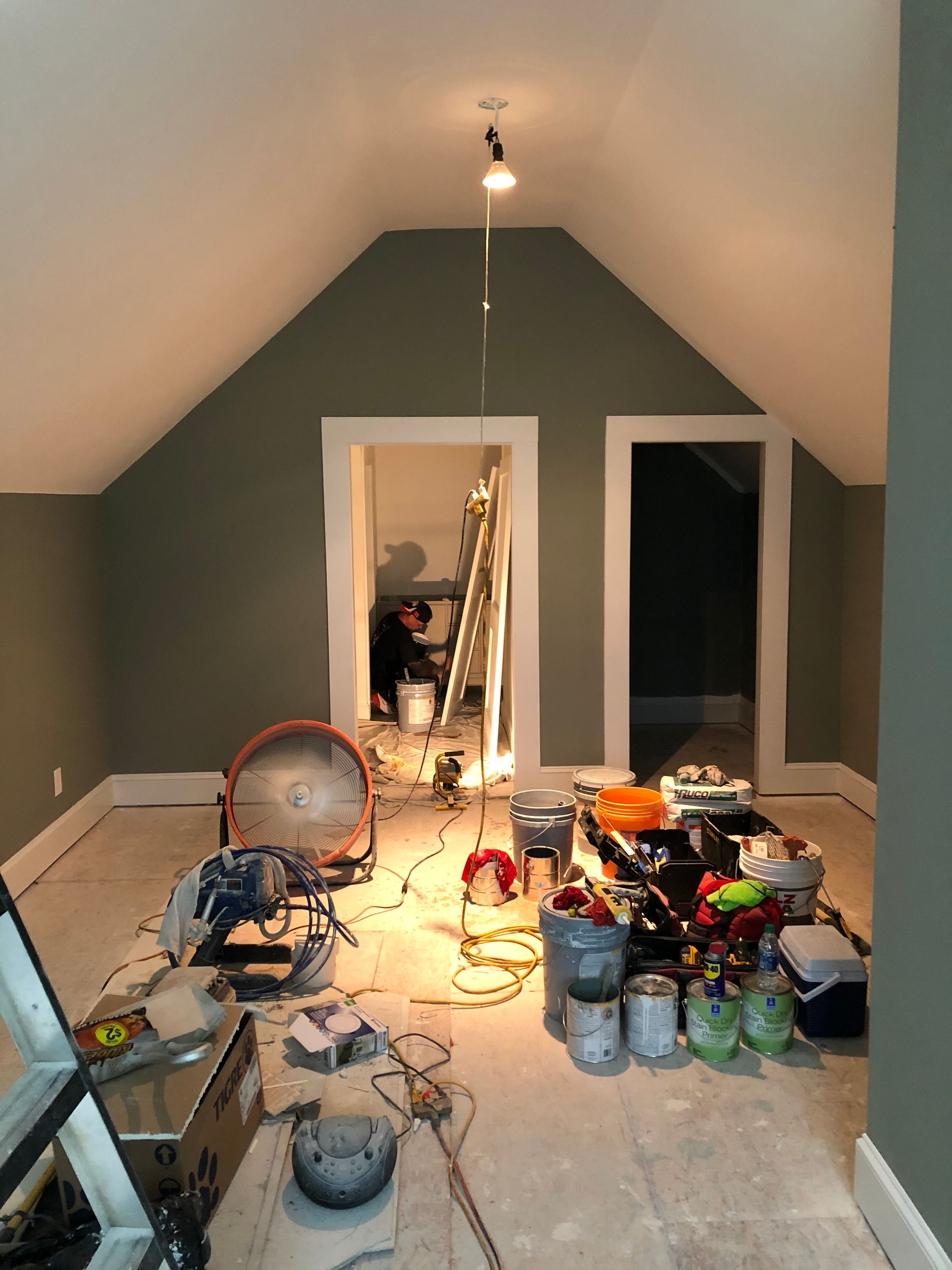 Unfinished attic room with green walls, cluttered floor, tools, cords, and paint cans under a single light.