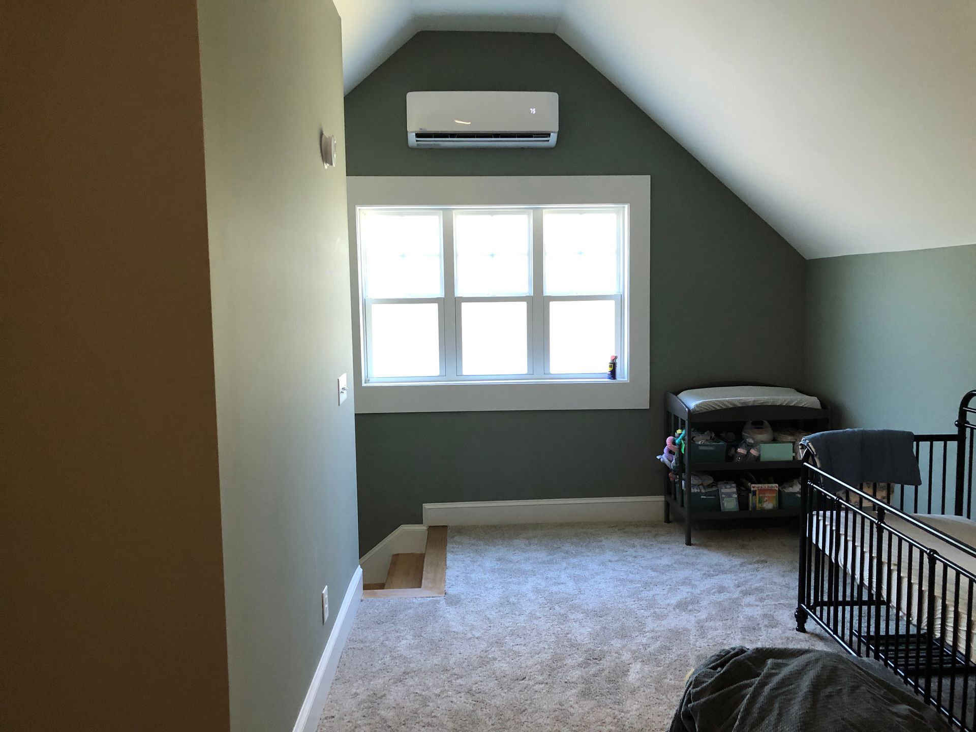 Slanted bedroom with gray walls, window AC, bright window, carpet, and black crib railing.