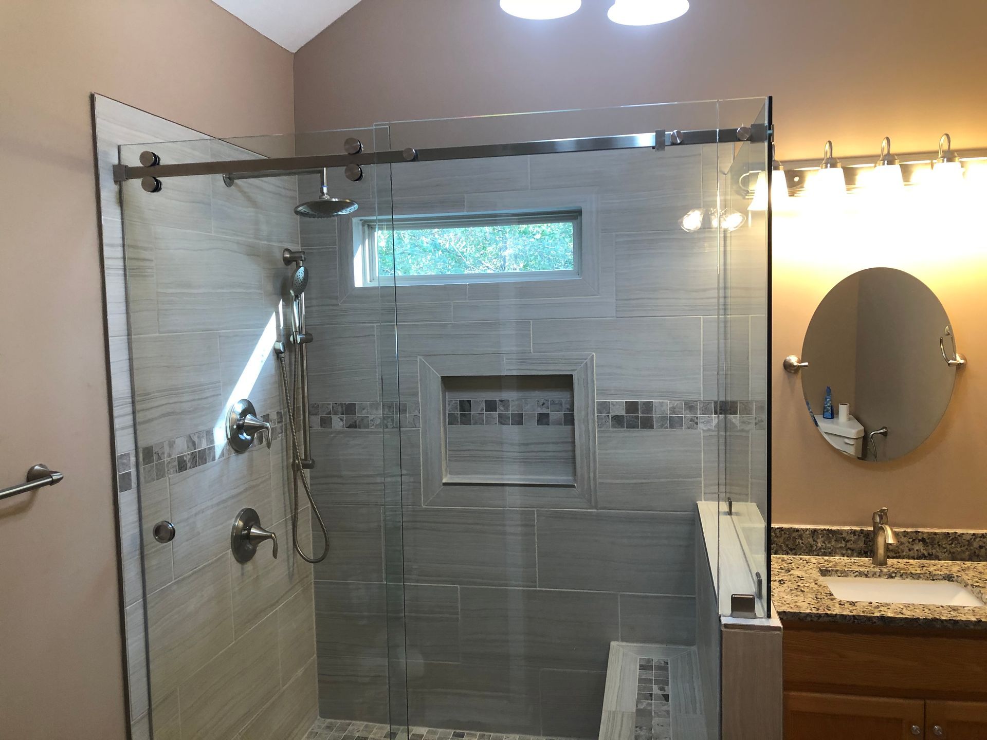Modern bathroom with glass shower doors, gray tile, and a vanity with round mirror under warm lights