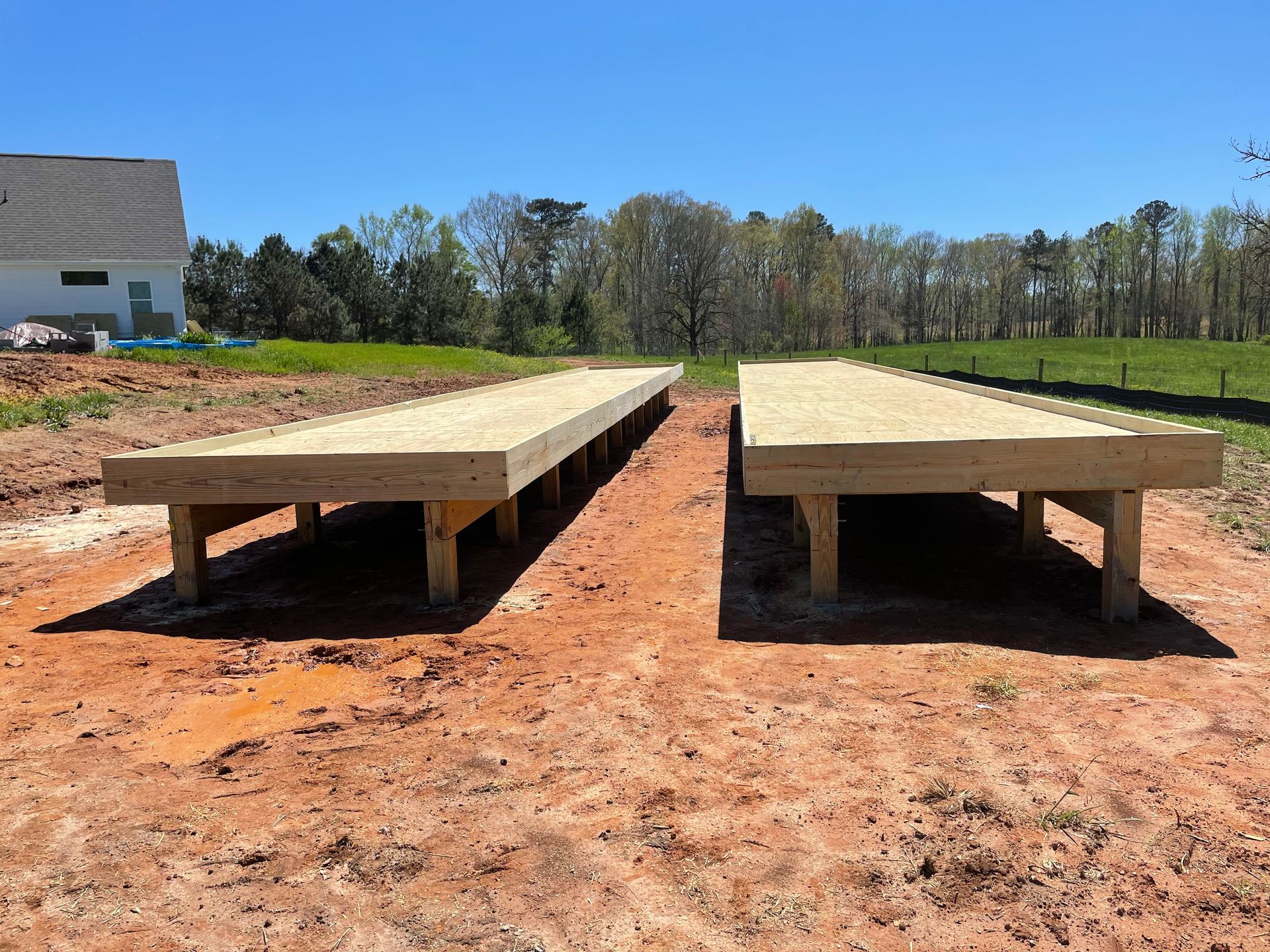 Two long wooden platforms on reddish dirt, beside a house and grassy field under a blue sky.