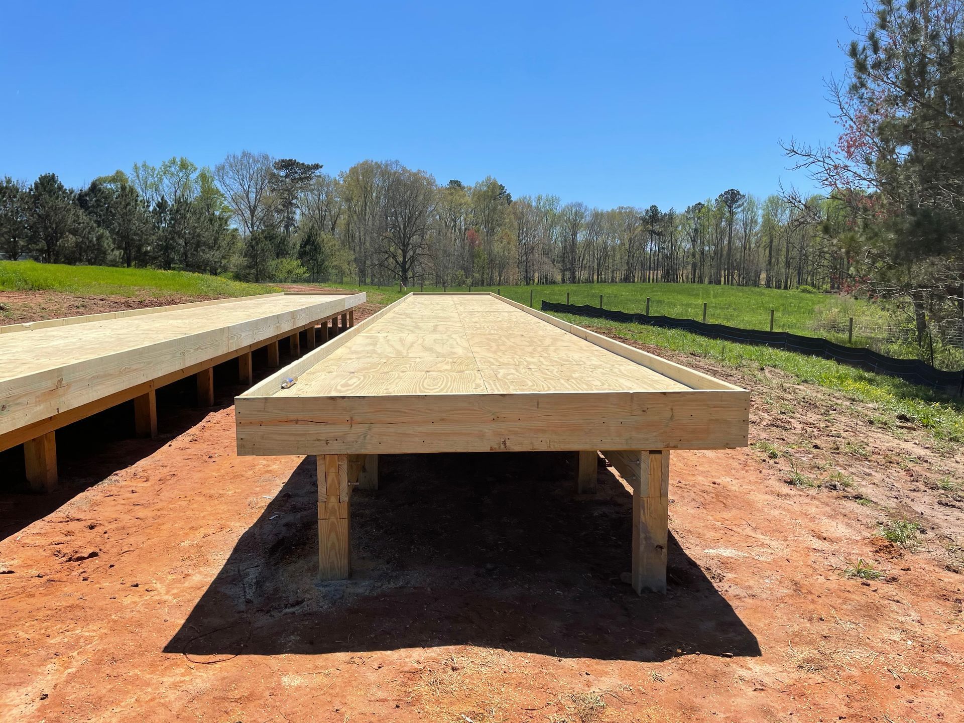 Wooden outdoor platform on supports in a sunny field, with trees and blue sky in the background