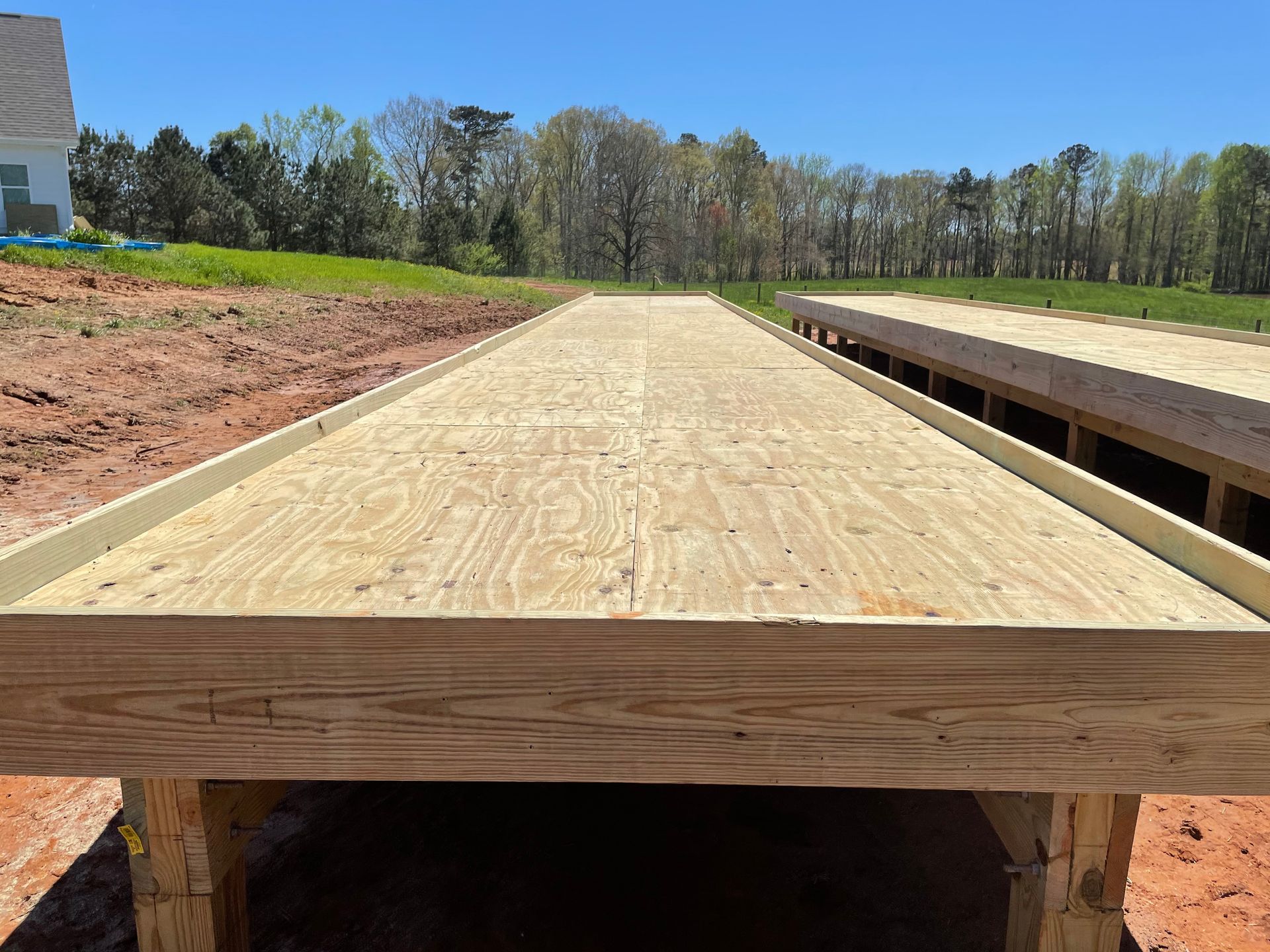 Wooden deck framing under construction beside a dirt lot and grassy field under a clear blue sky