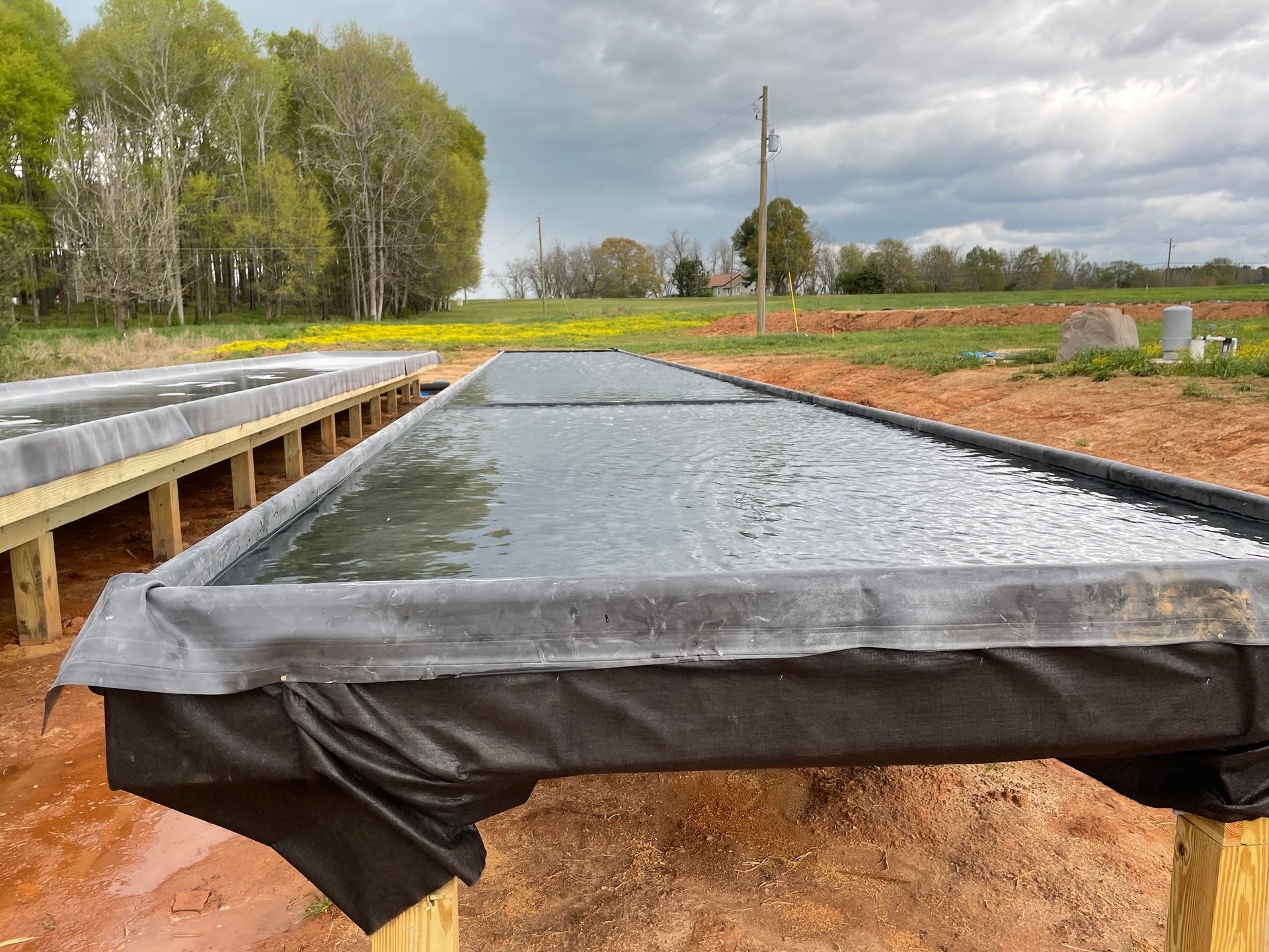 Rectangular black-lined pond or raised bed filled with water in a muddy field under cloudy sky