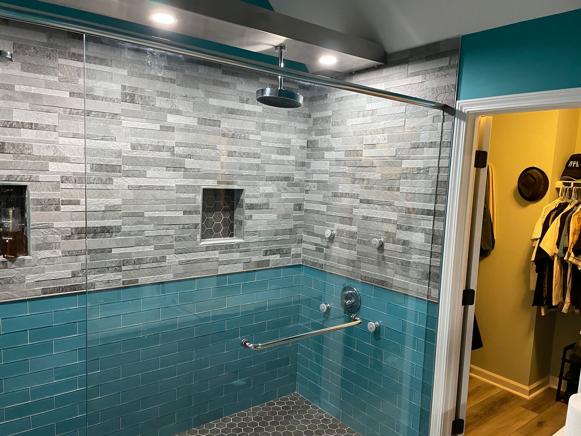 Blue-tiled shower with gray stone wall, rainfall showerhead, and open doorway to a closet.
