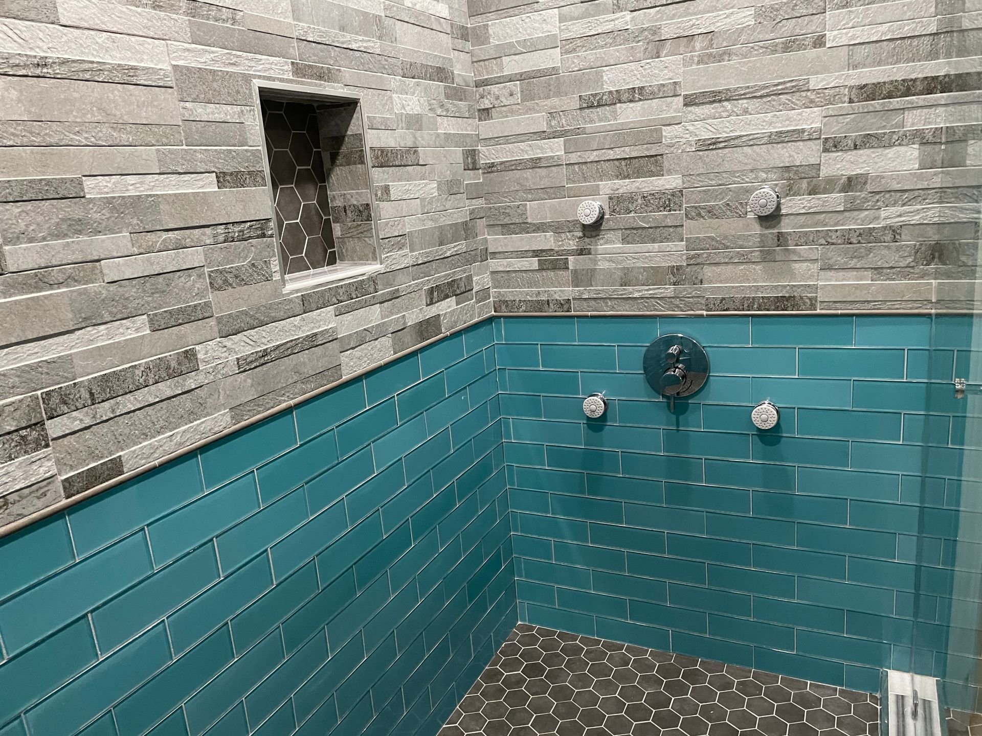 Small tiled bathroom shower with teal lower walls and gray stone-pattern upper walls.