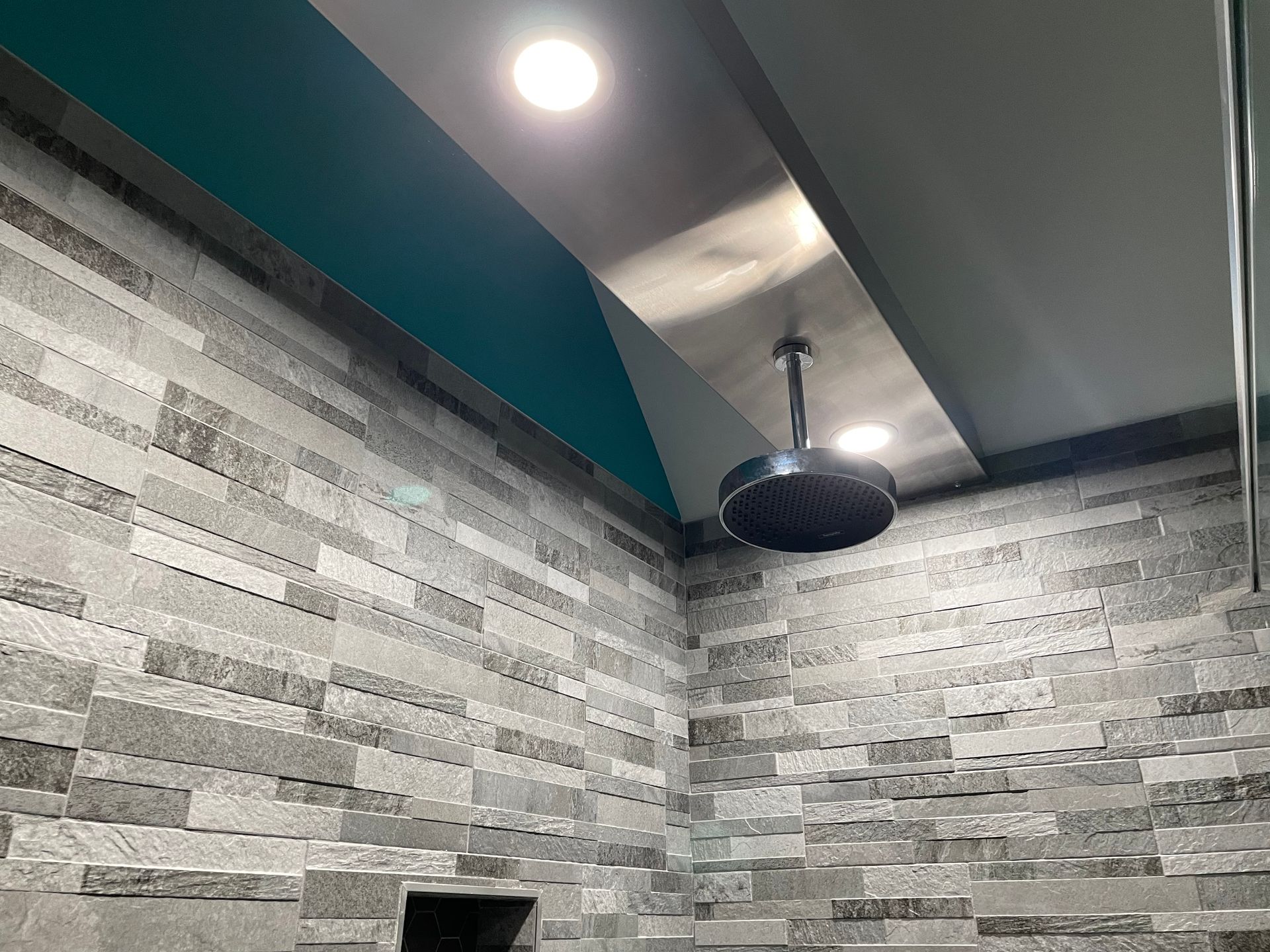 Modern bathroom corner with gray stone walls, teal accent trim, ceiling lights, and a black rain showerhead