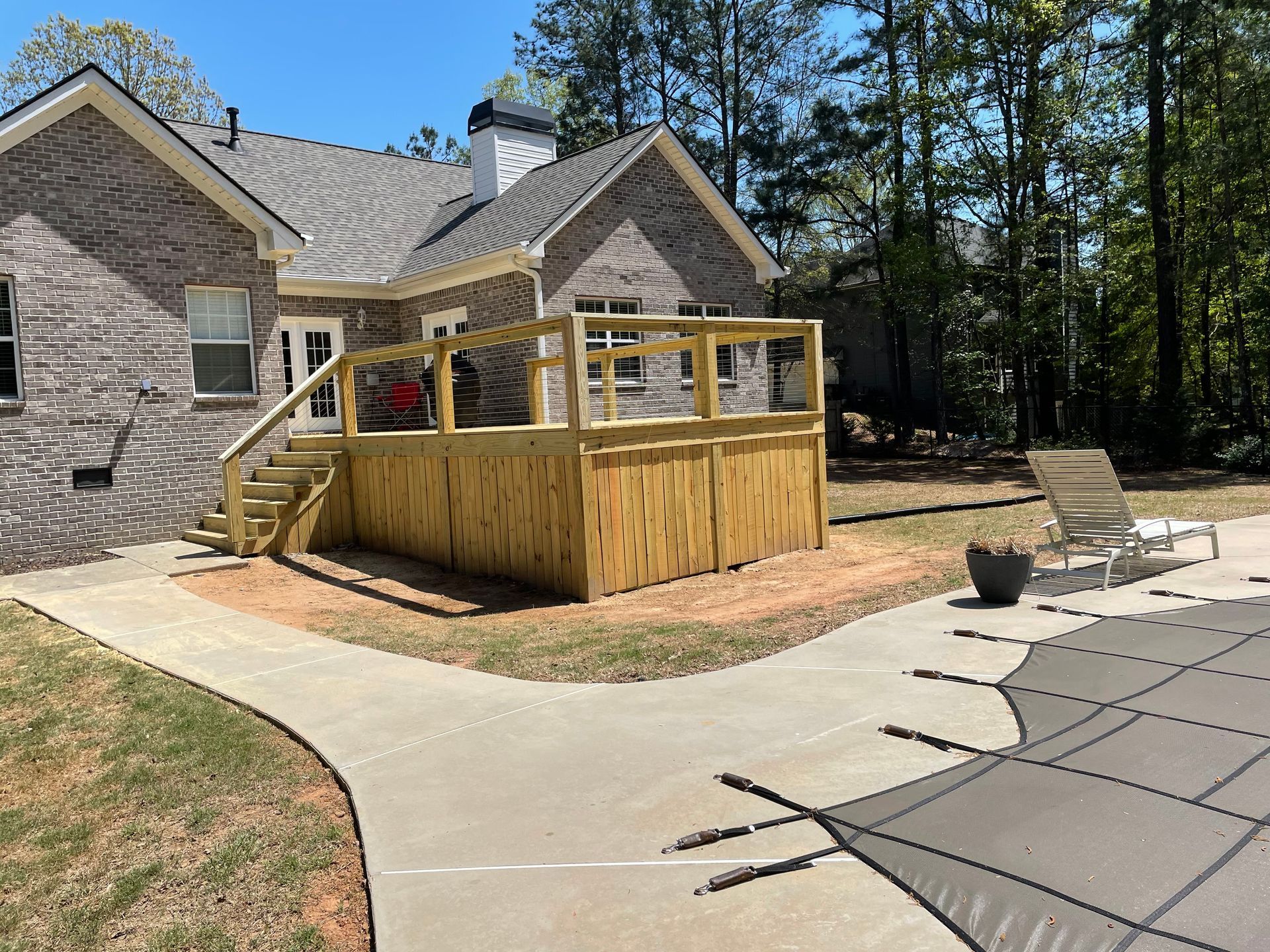 Stone house with a wooden deck and ramp beside a curved concrete patio, surrounded by trees.