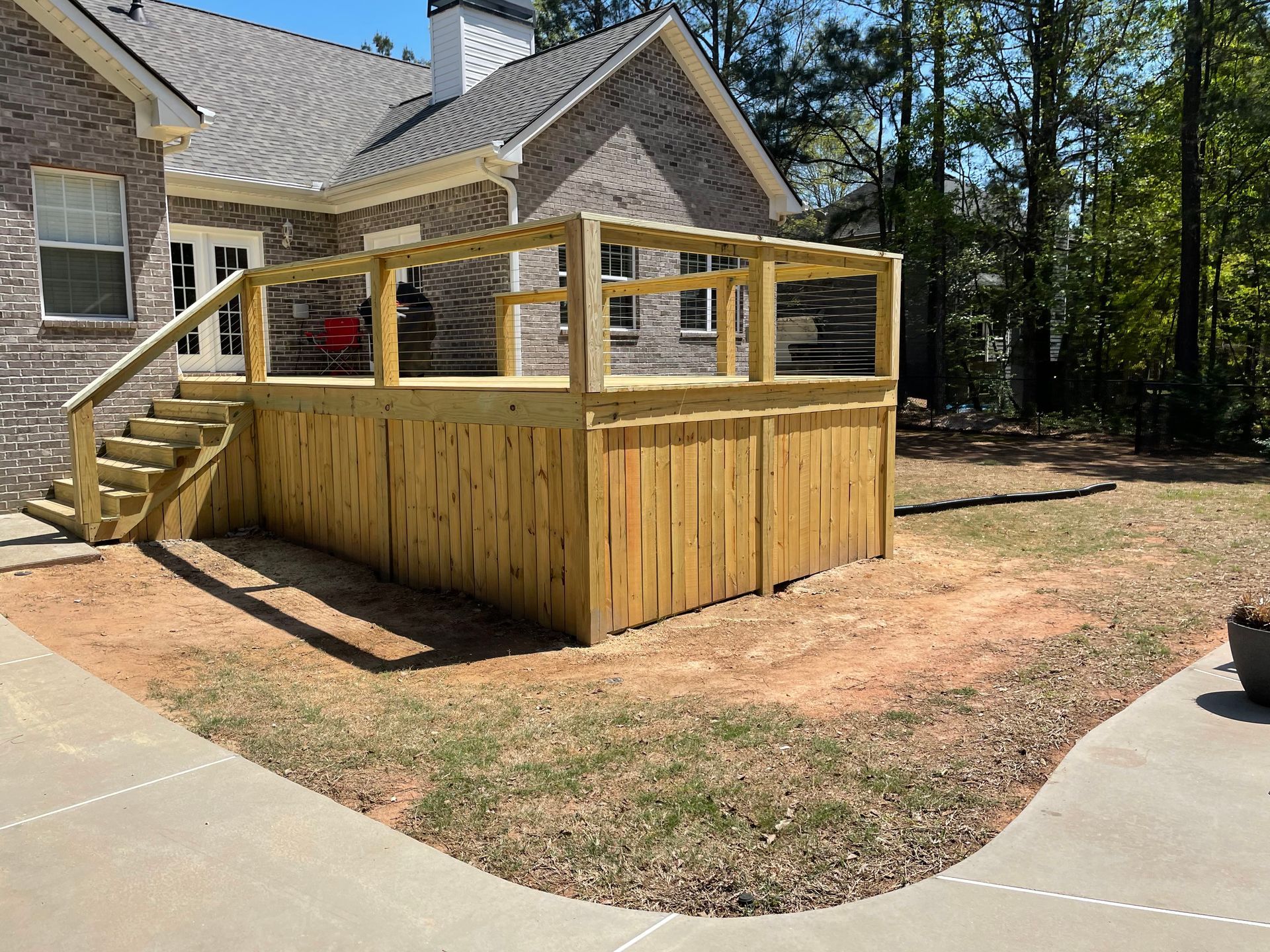 Raised wooden deck with railing beside a house, surrounded by trees and a curved concrete driveway.