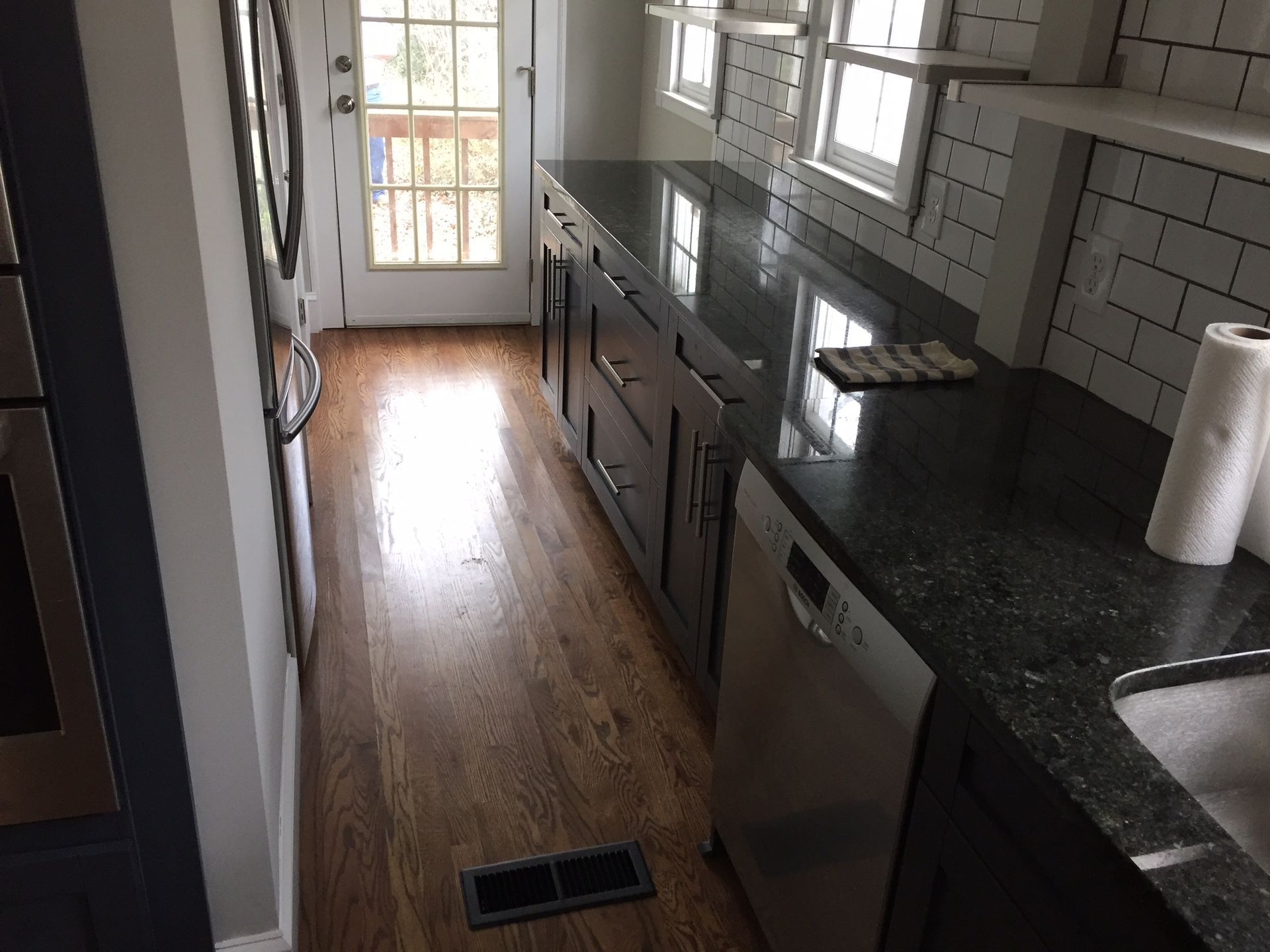 Narrow kitchen with dark counters, light cabinets, wood floor, and a bright door at the end.