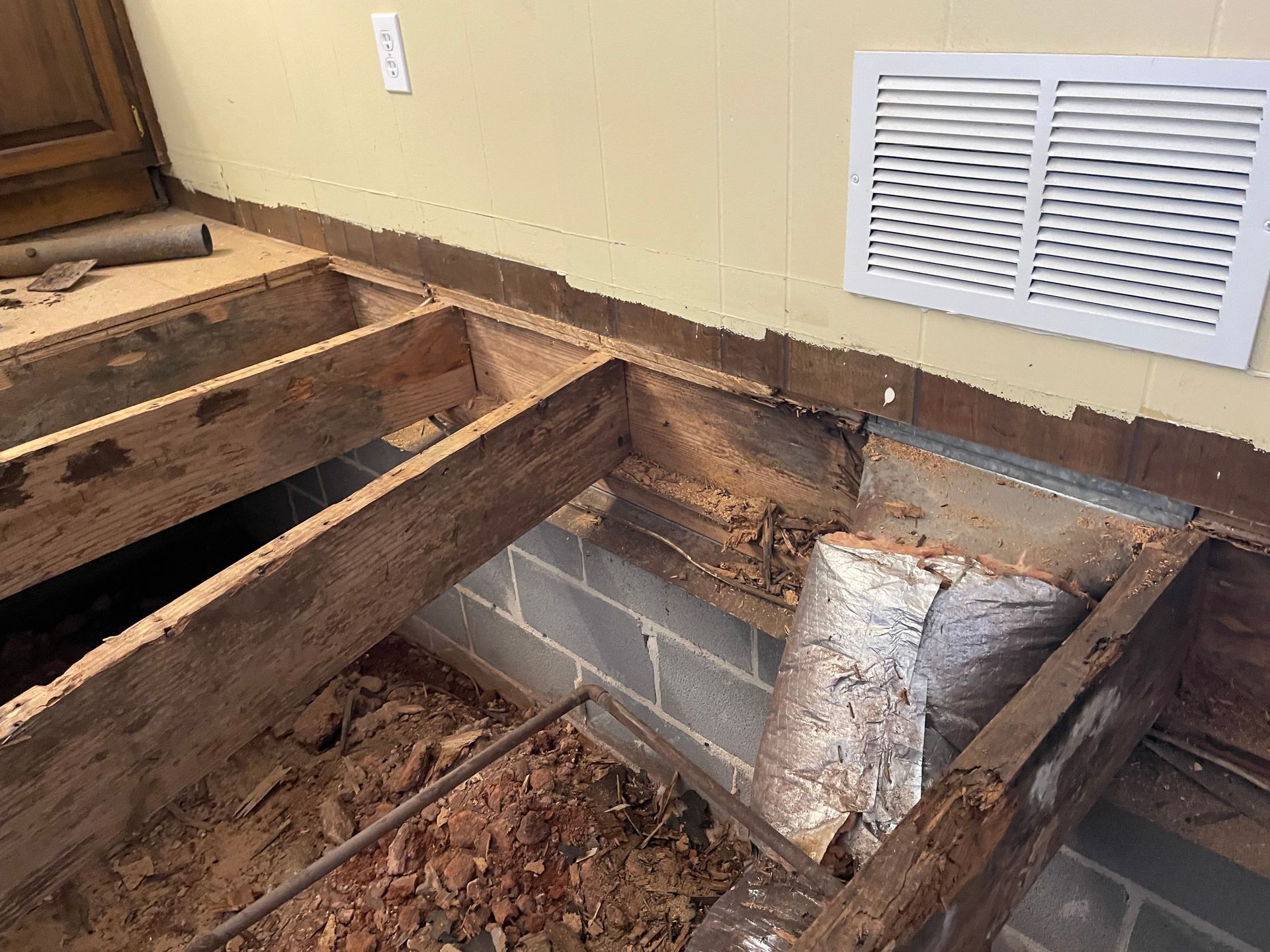 Damaged floor with exposed joists and debris beside a wall vent and wooden steps