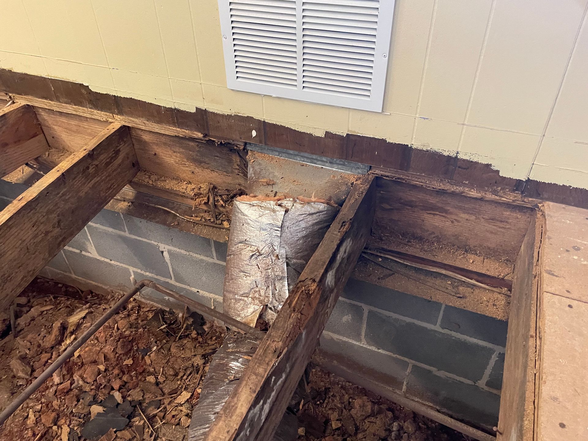 Exposed floor joists and cinder blocks beneath a damaged porch deck near a house vent.