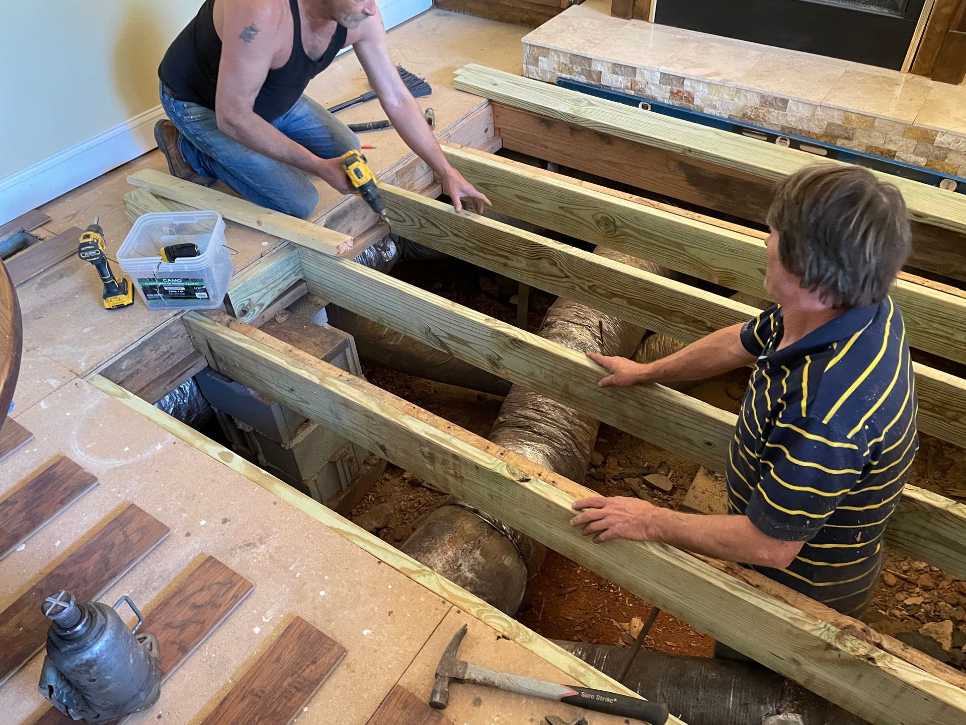 Two people installing wooden floor joists in a room with tools and exposed subfloor.