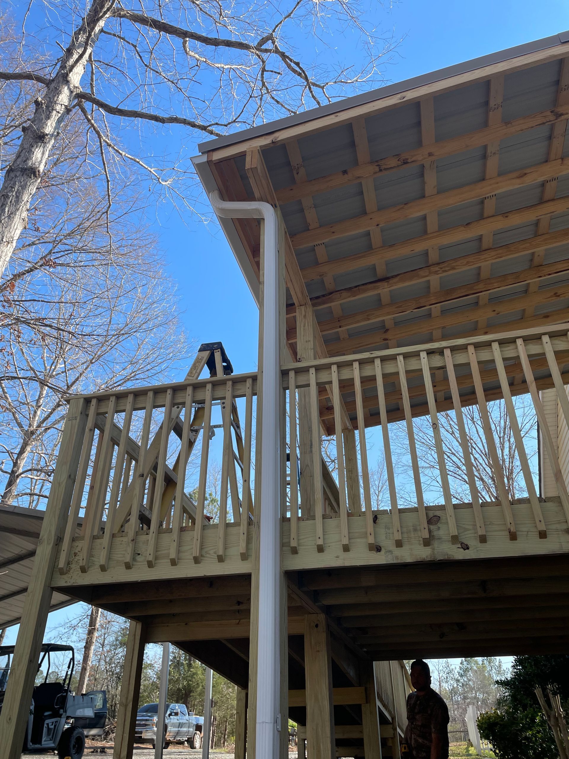 Wooden deck with railings beneath a covered roof, viewed from below against a clear blue sky.
