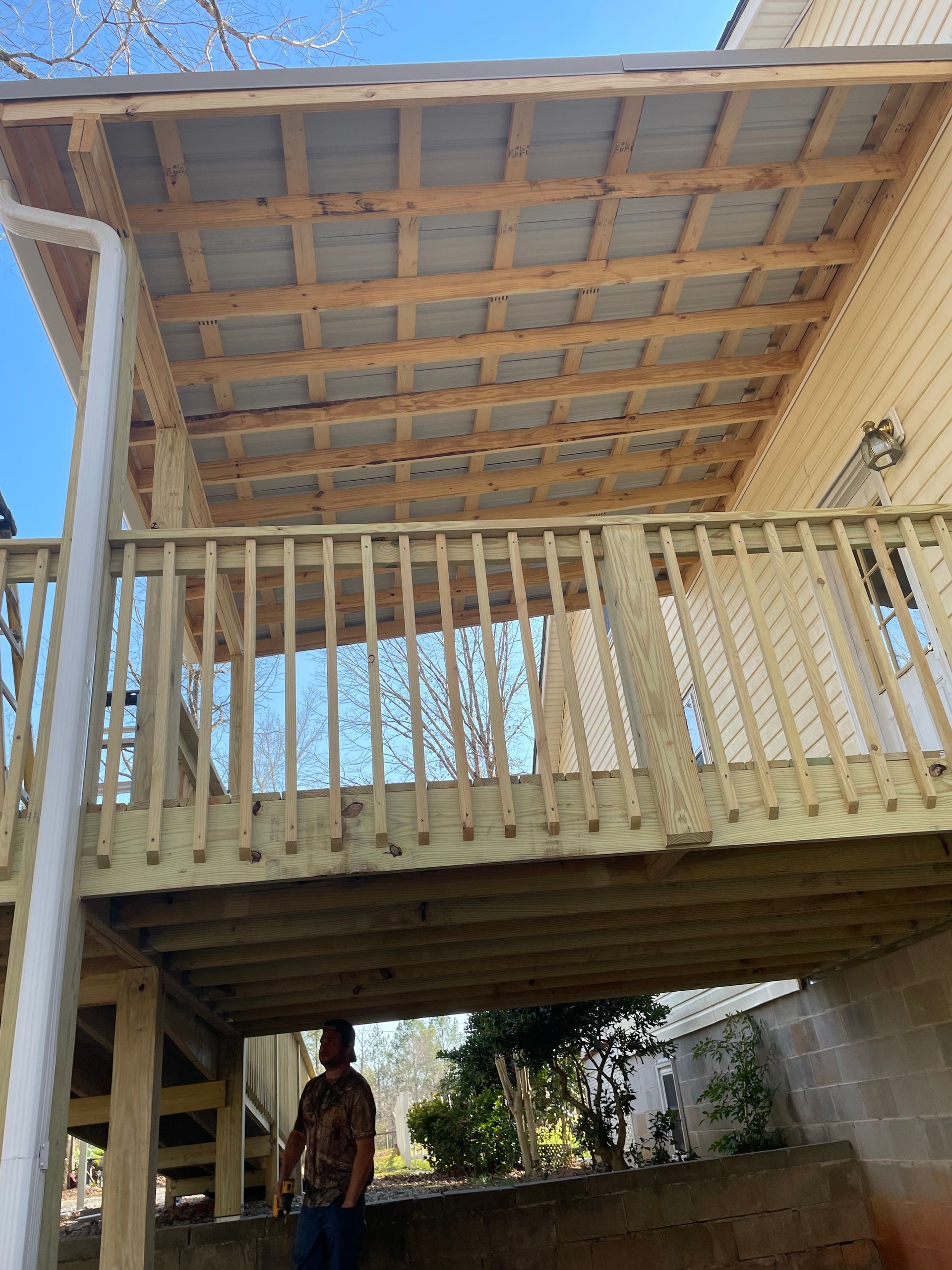 Covered wooden deck with railing under a corrugated roof; person standing beneath in the shaded area.