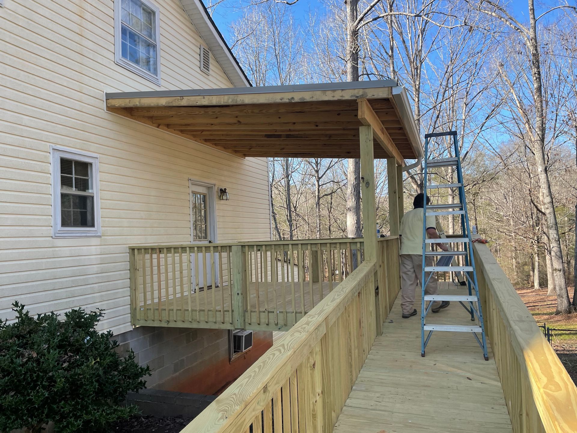 Covered wooden ramp under construction beside a beige house, with a ladder and leafless trees.