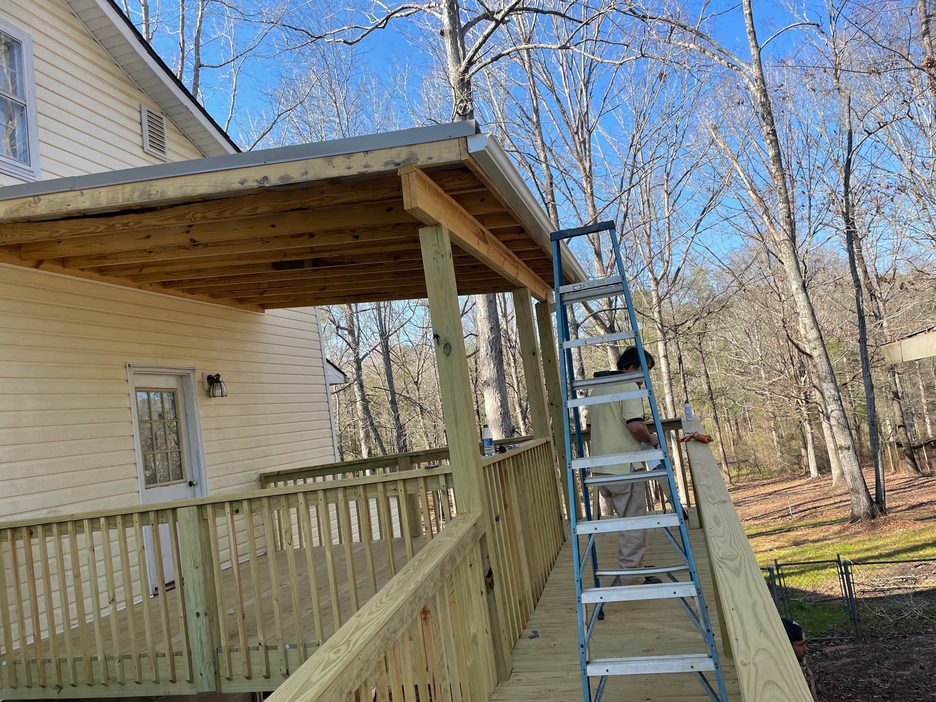 Porch with wooden railing and roof beside a house, with a ladder leaning against it and trees in the background
