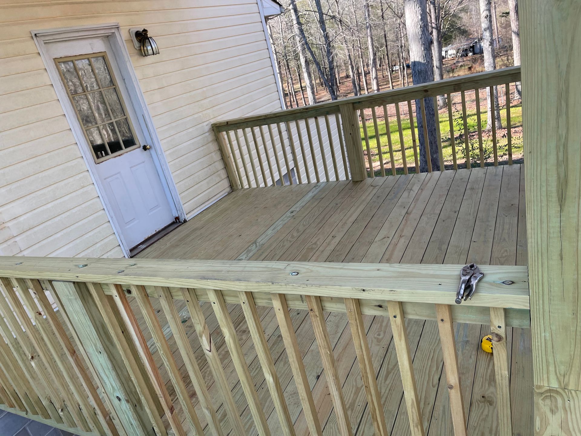 Wooden deck with railings beside a white house and door, overlooking a yard and trees.