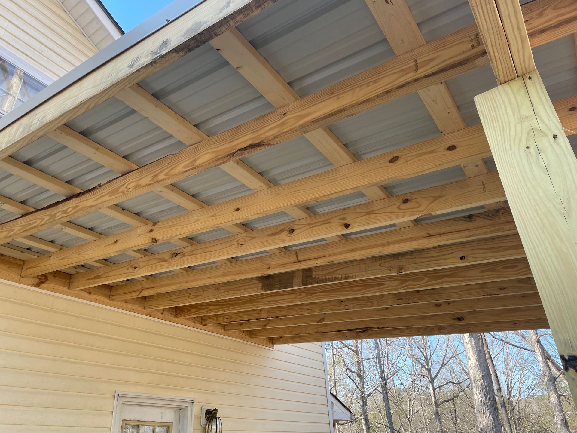 Wooden deck roof framing with exposed beams under a house eave, viewed from below