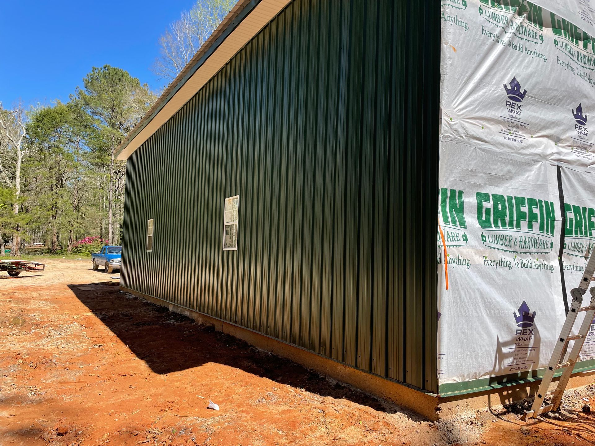 Long green metal building with white wrapped end wall on a wooded dirt lot under blue sky