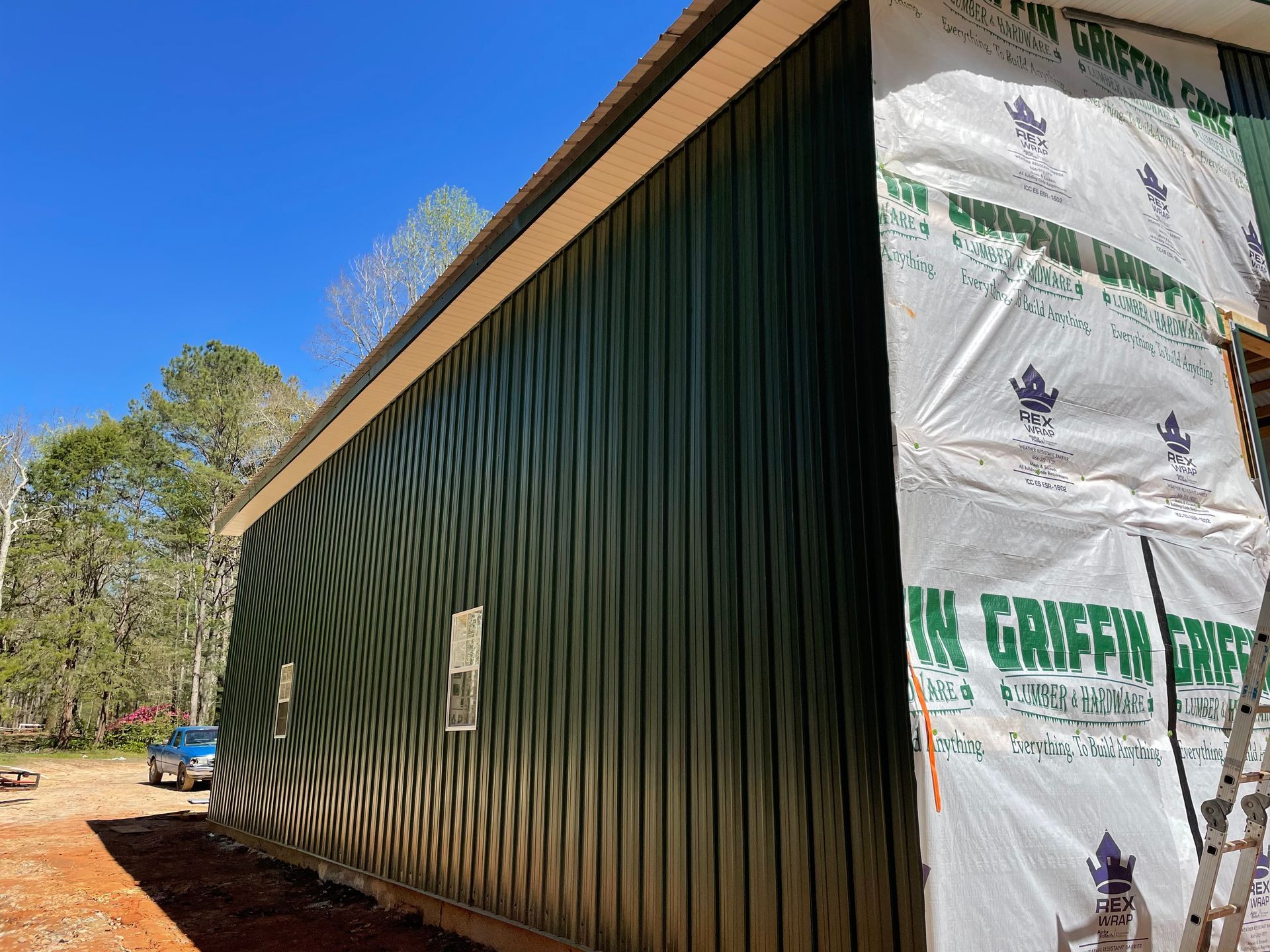 Green-sided shed under construction with white wrapped end, outdoors against blue sky