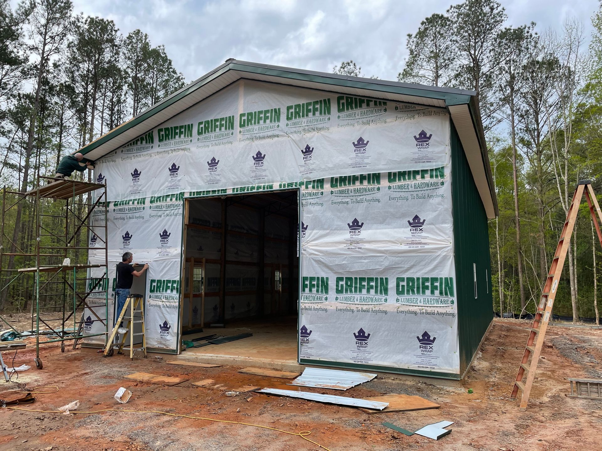 Partially built house wrapped in white house wrap at a wooded construction site, with a worker nearby