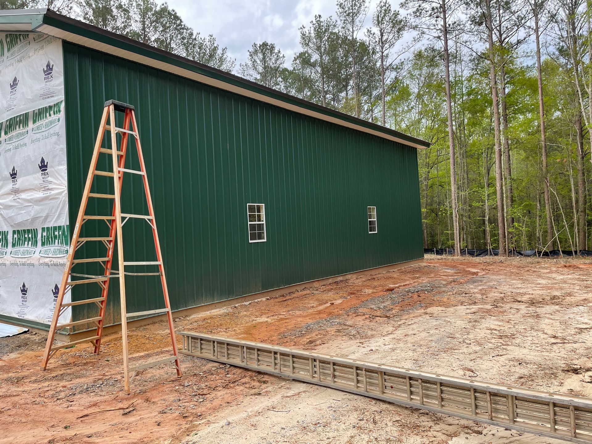Green metal building under construction with a ladder and concrete foundation, surrounded by trees