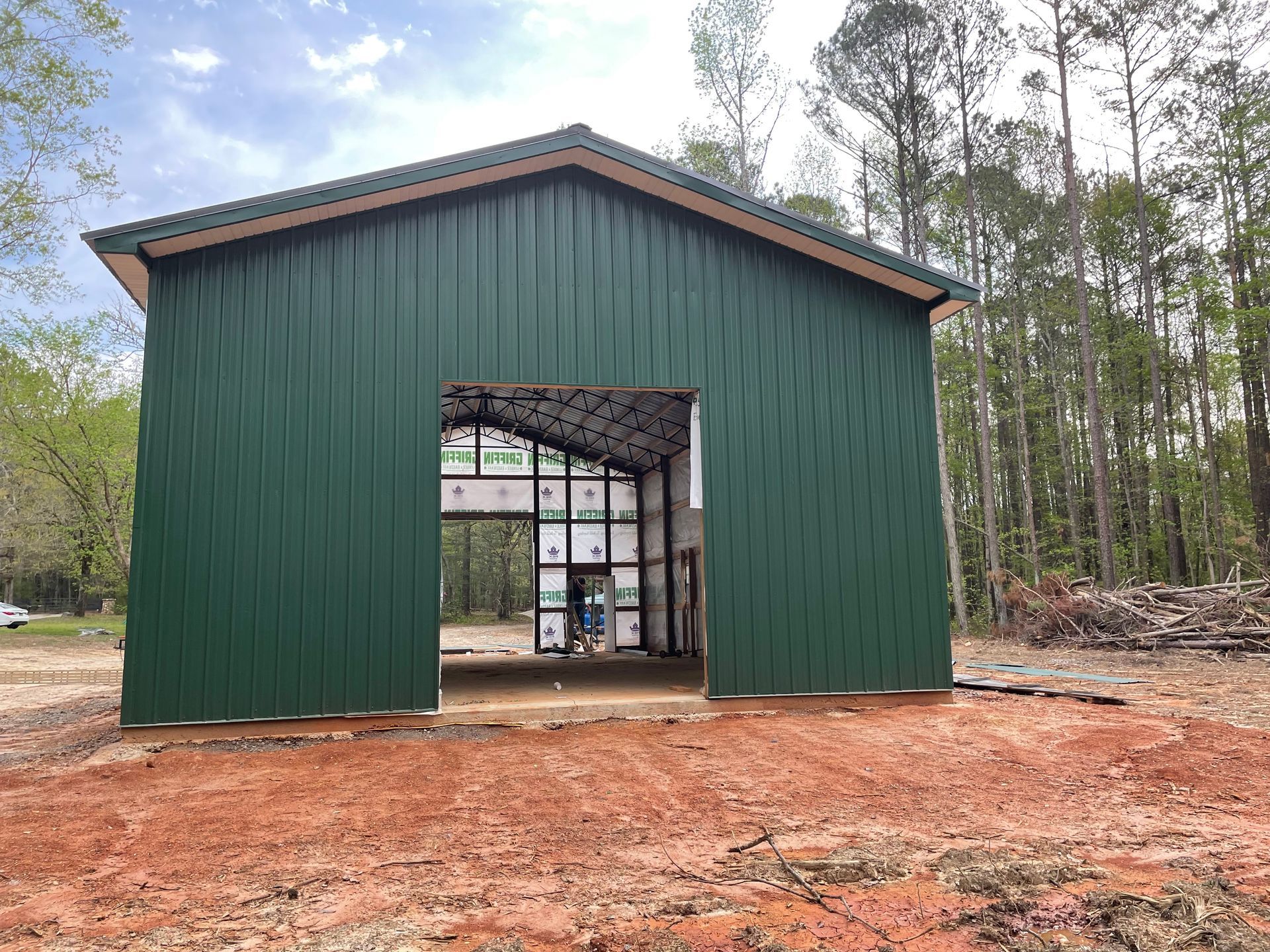Green metal shed with open front on a red dirt lot, backed by trees
