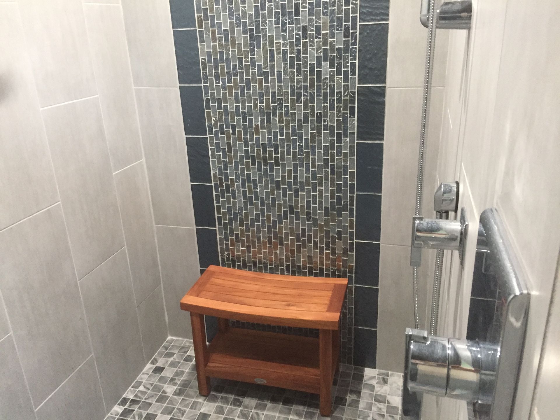 Small tiled shower with a wooden bench and chrome grab bars.