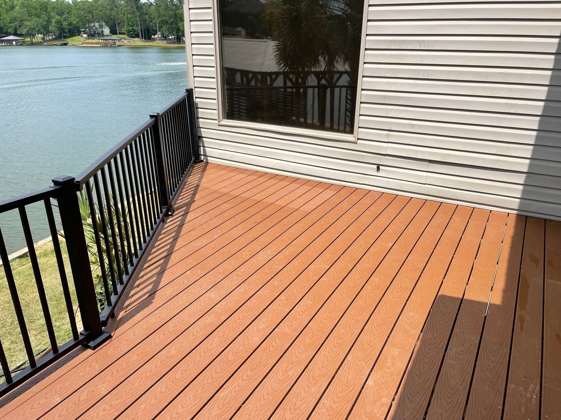 Wooden deck beside a lake with black railing and house siding, sunlight casting shadows