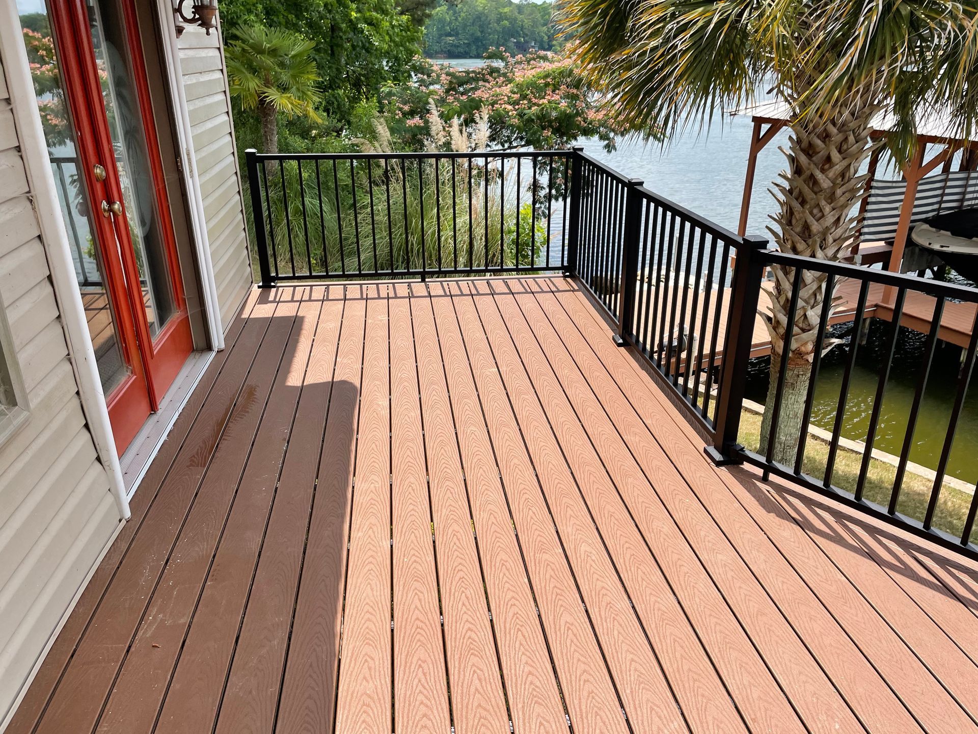 Wooden deck with black railings overlooking a lake and trees, beside a house with red-trimmed doors.