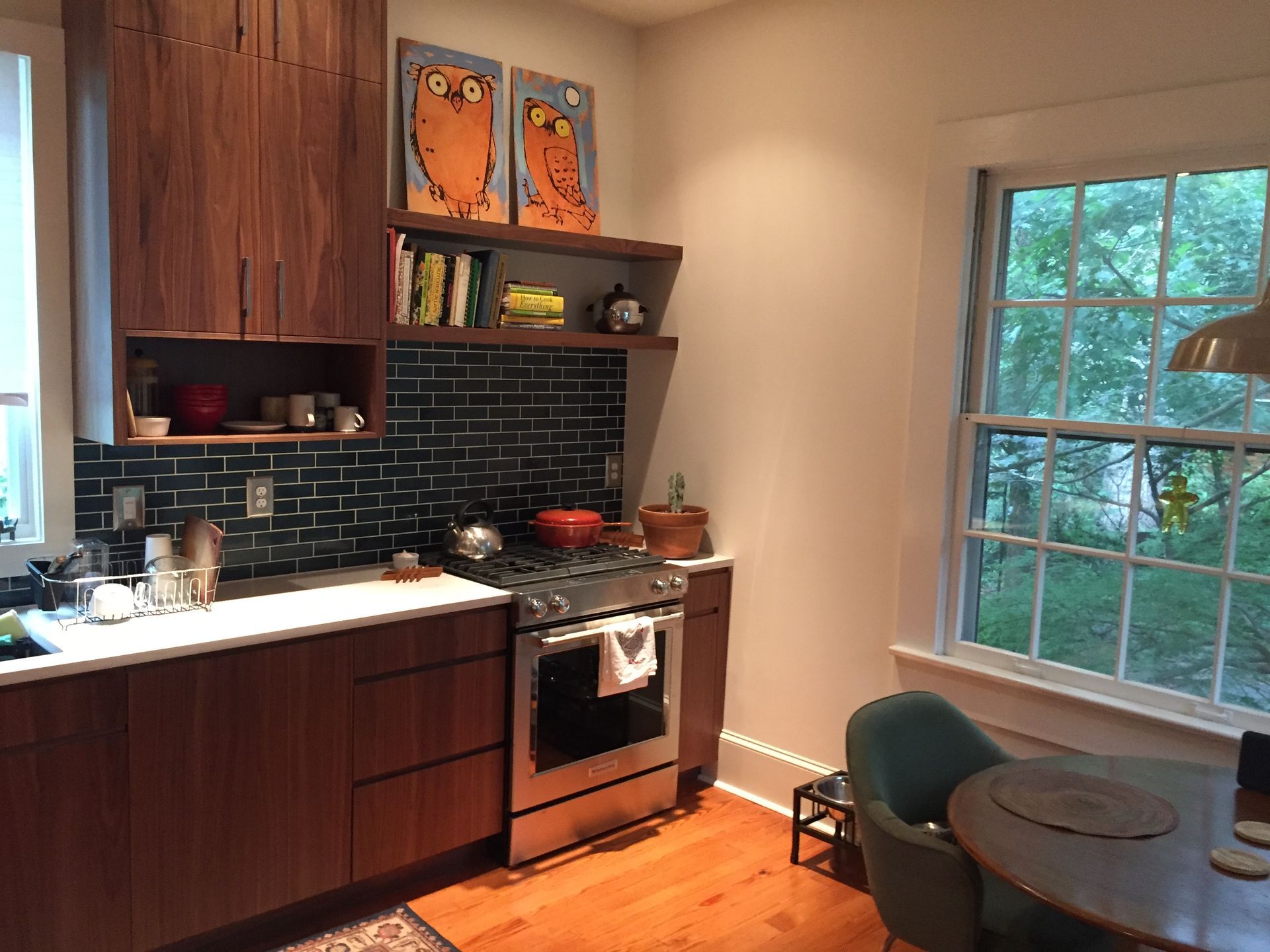 Small kitchen with dark cabinets, black backsplash, stove, sink, and a round table by a bright window.